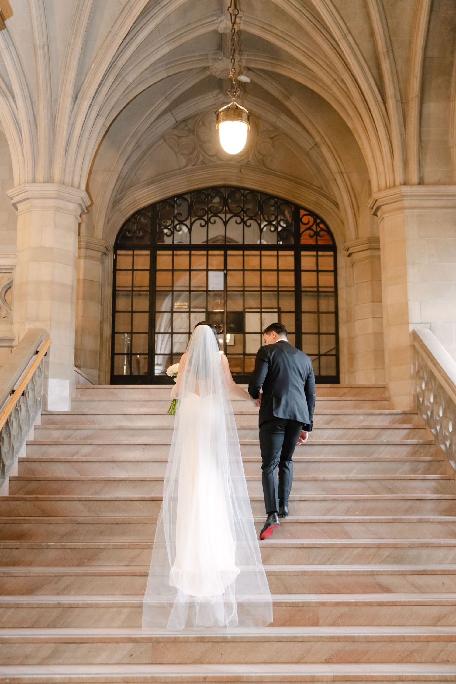 Long flowing veil trails behind as couple ascends grand marble staircase under vaulted ceiling.