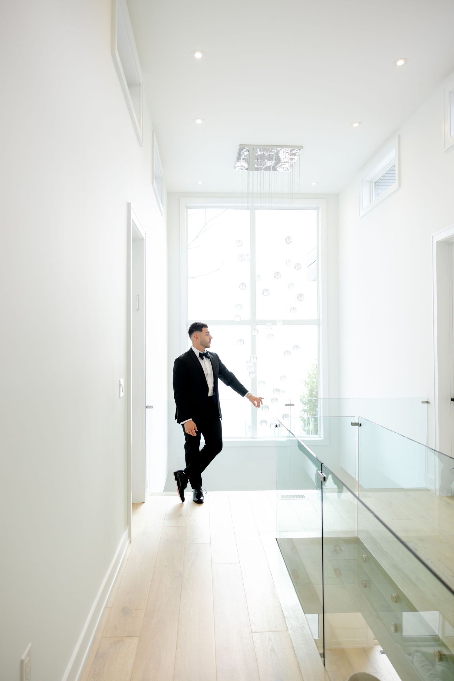 Groom in classic tuxedo walks down bright modern hallway with glass railings and wooden floors.