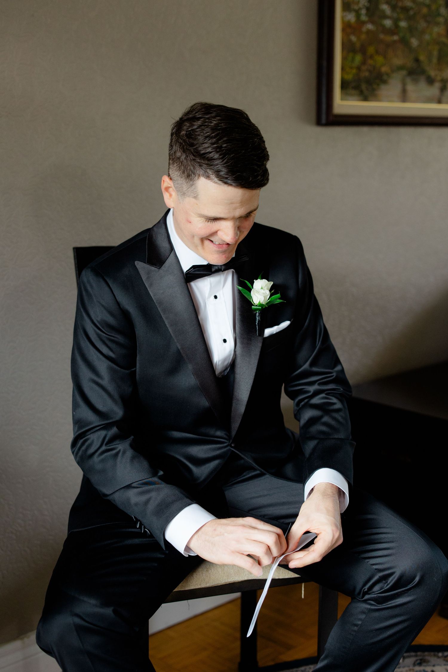 Wedding day portrait showing someone in a black tuxedo with white boutonniere seated indoors.