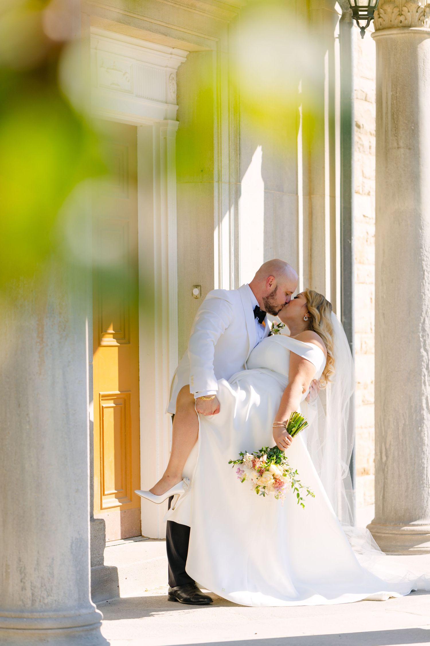 Romantic wedding pose between white columns with soft natural lighting and bokeh effects.
