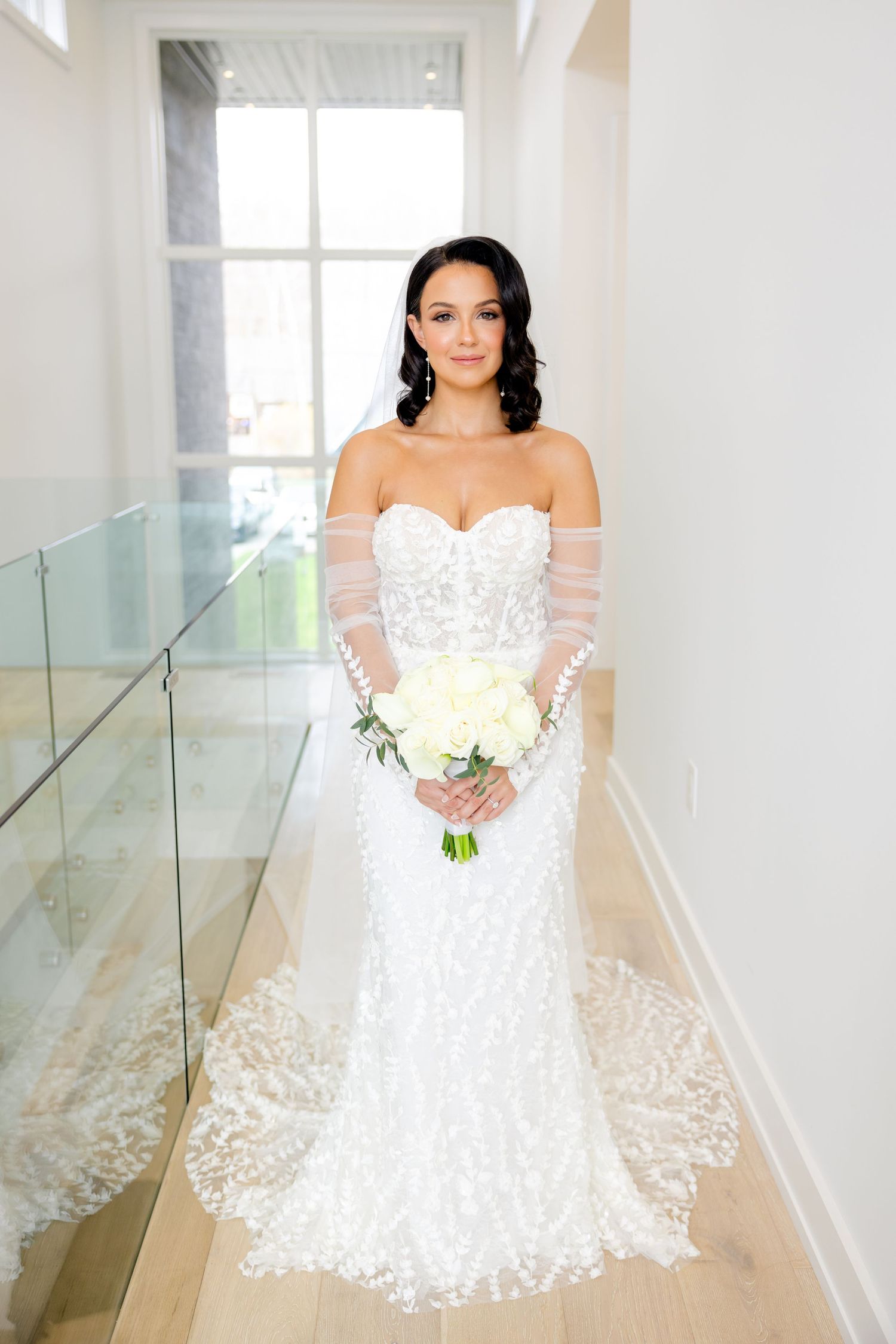 A stunning bridal portrait showing an elegant lace wedding gown with sheer sleeves and a white bouquet in a bright hallway.