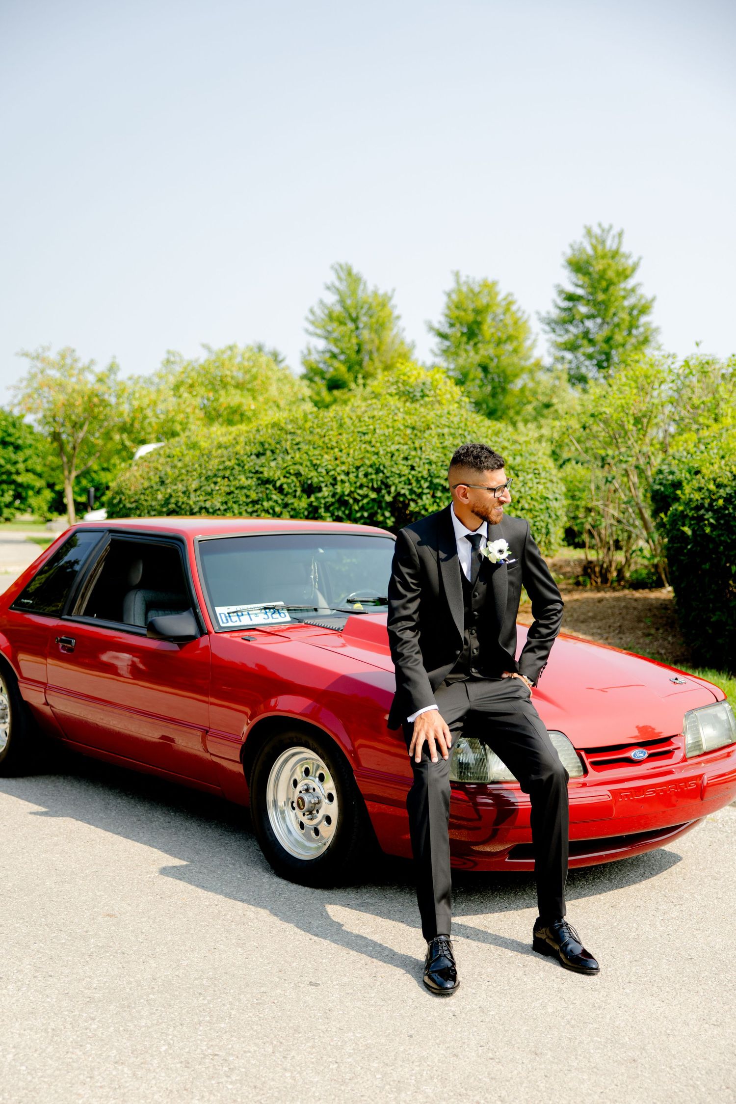 Groom in formal black suit sits casually on hood of vintage red Mustang car.