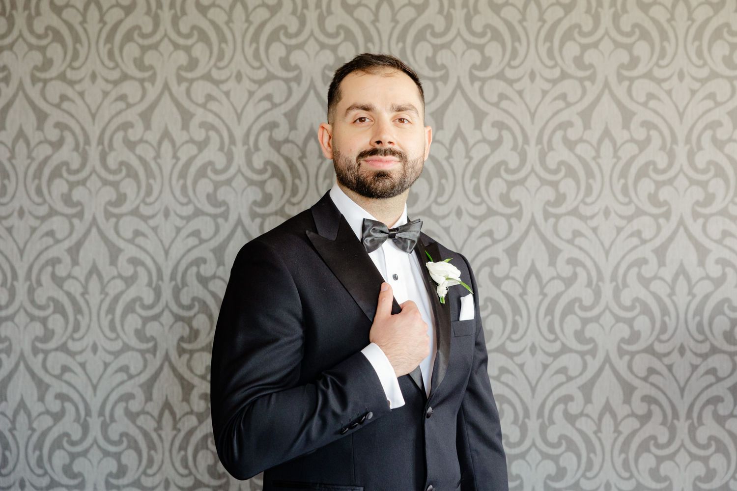 A close-up portrait of someone adjusting their bowtie in a black tuxedo against grey wallpaper.