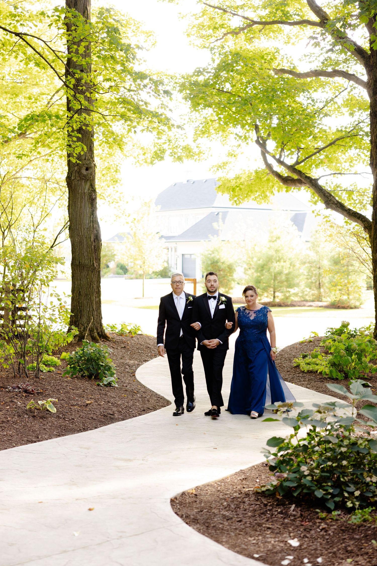A winding garden path with formal-dressed attendees walking towards a building in the distance.