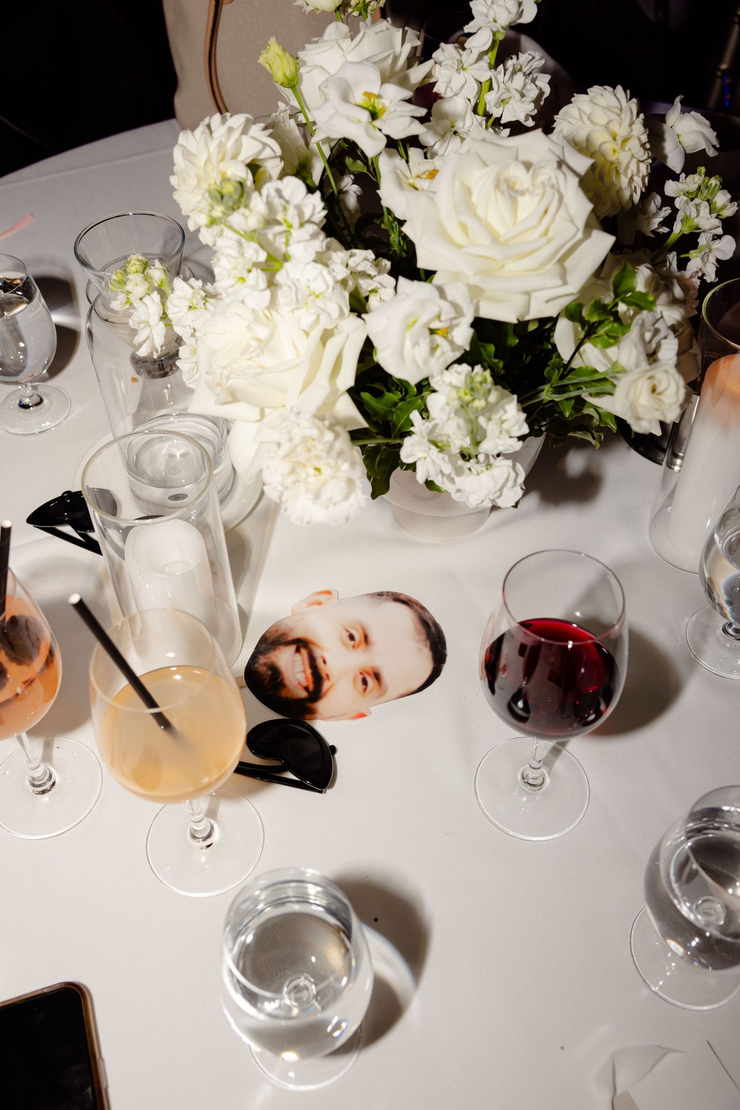 Elegant white floral centerpiece with wine glasses and decorative elements on wedding reception table.