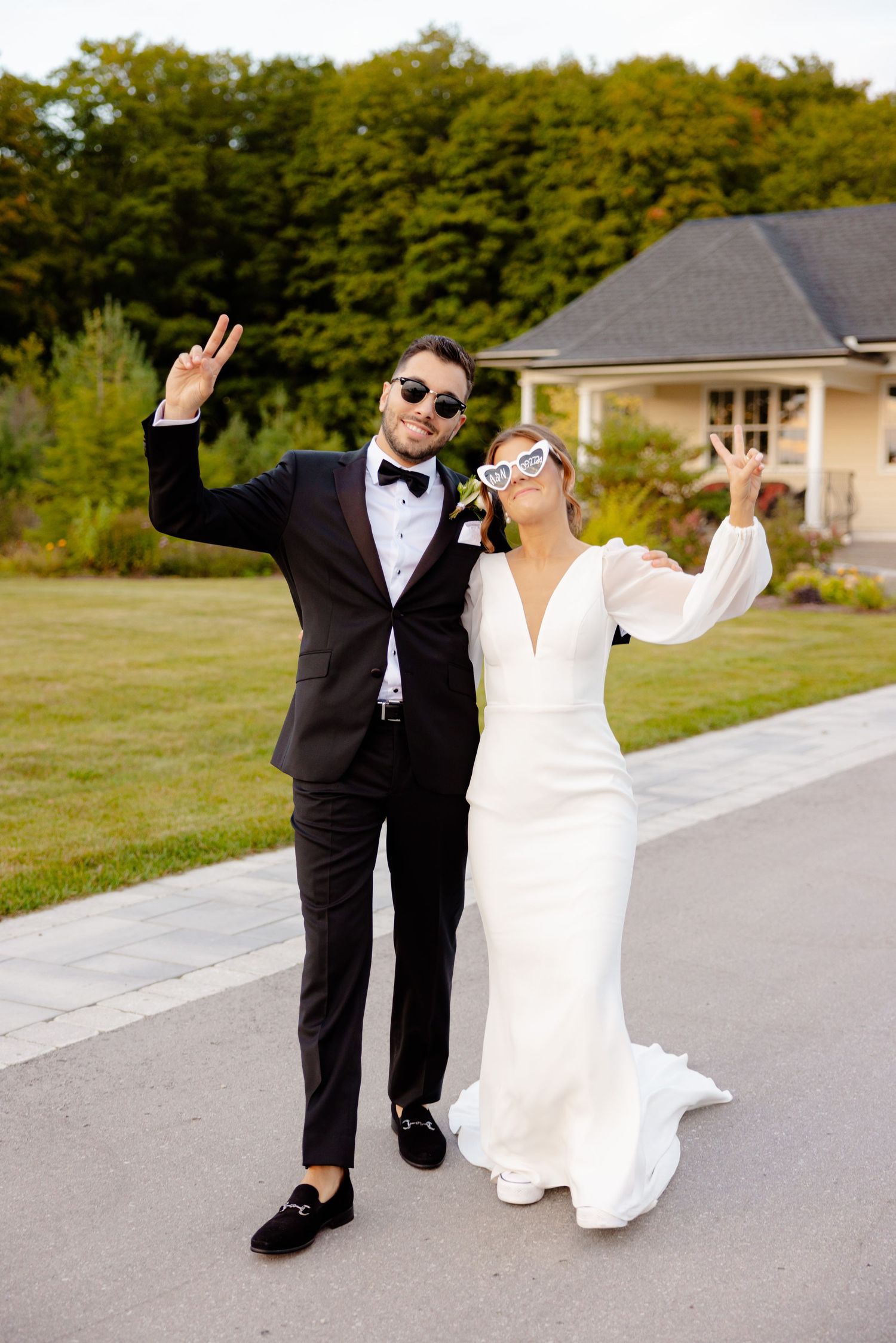 A playful wedding couple poses with peace signs and sunglasses on a paved path.