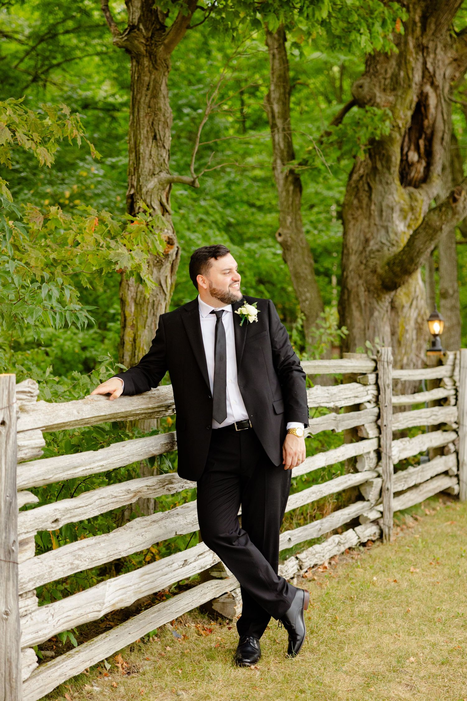 Groom in dark suit leans against rustic fence in garden setting.