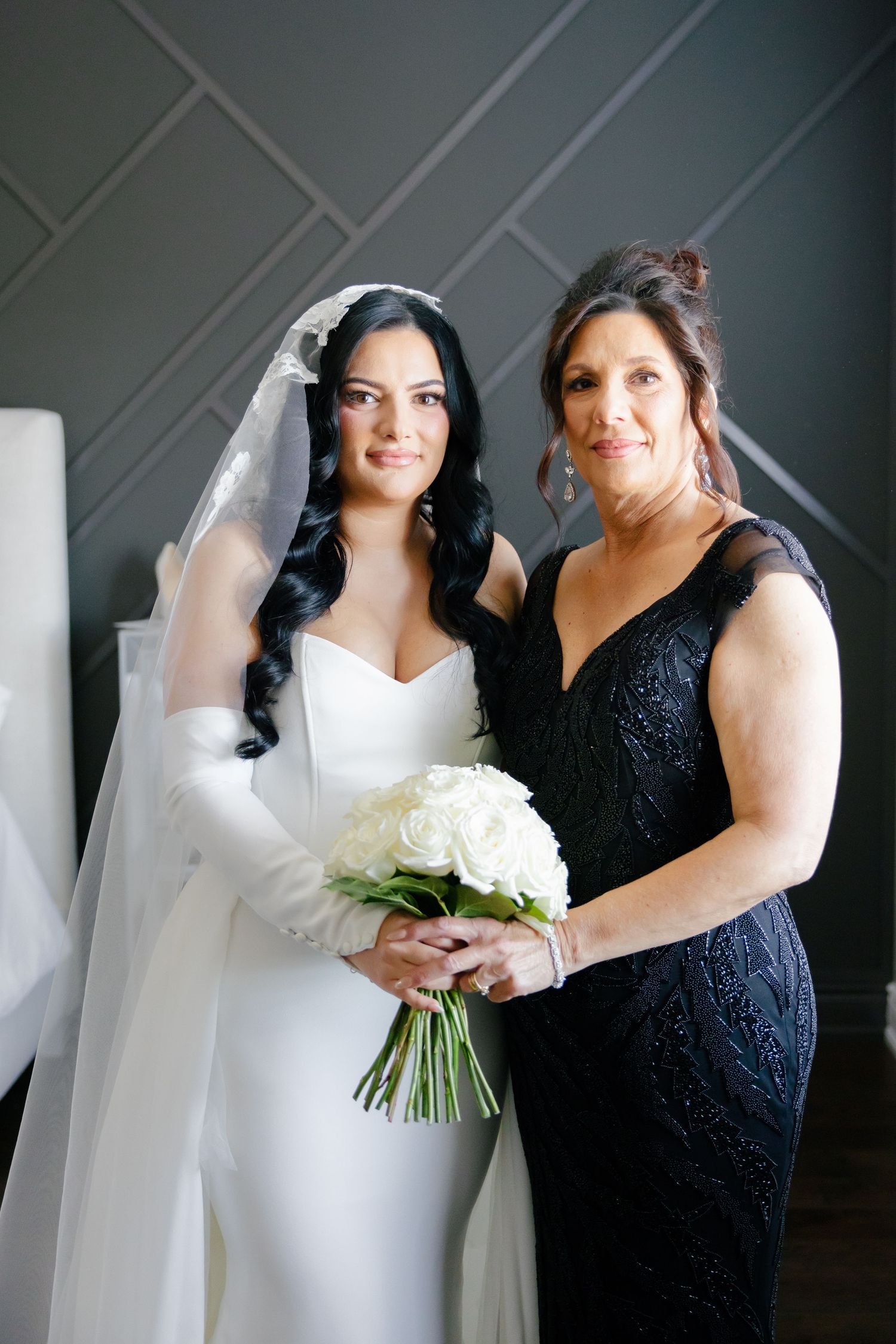 A bride in white holds a bouquet of roses while posing with a woman in a navy lace dress.