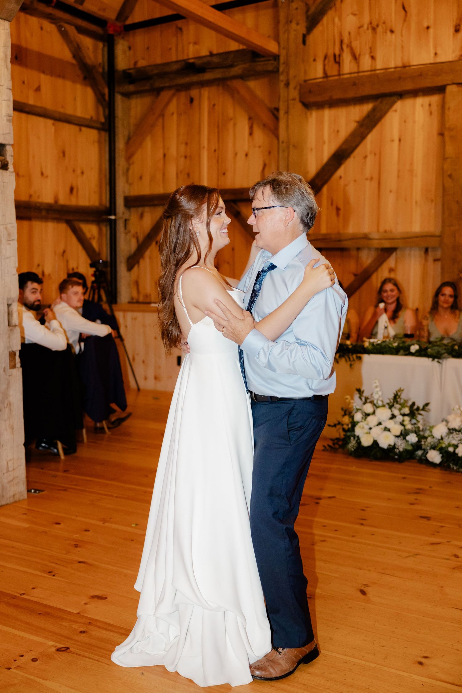 A couple shares a dance in a rustic barn venue decorated with white floral arrangements.
