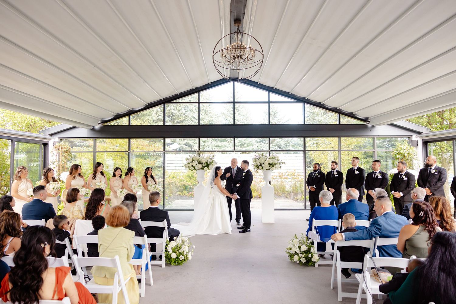 Bright indoor wedding ceremony with cathedral ceiling and guests seated on white folding chairs.