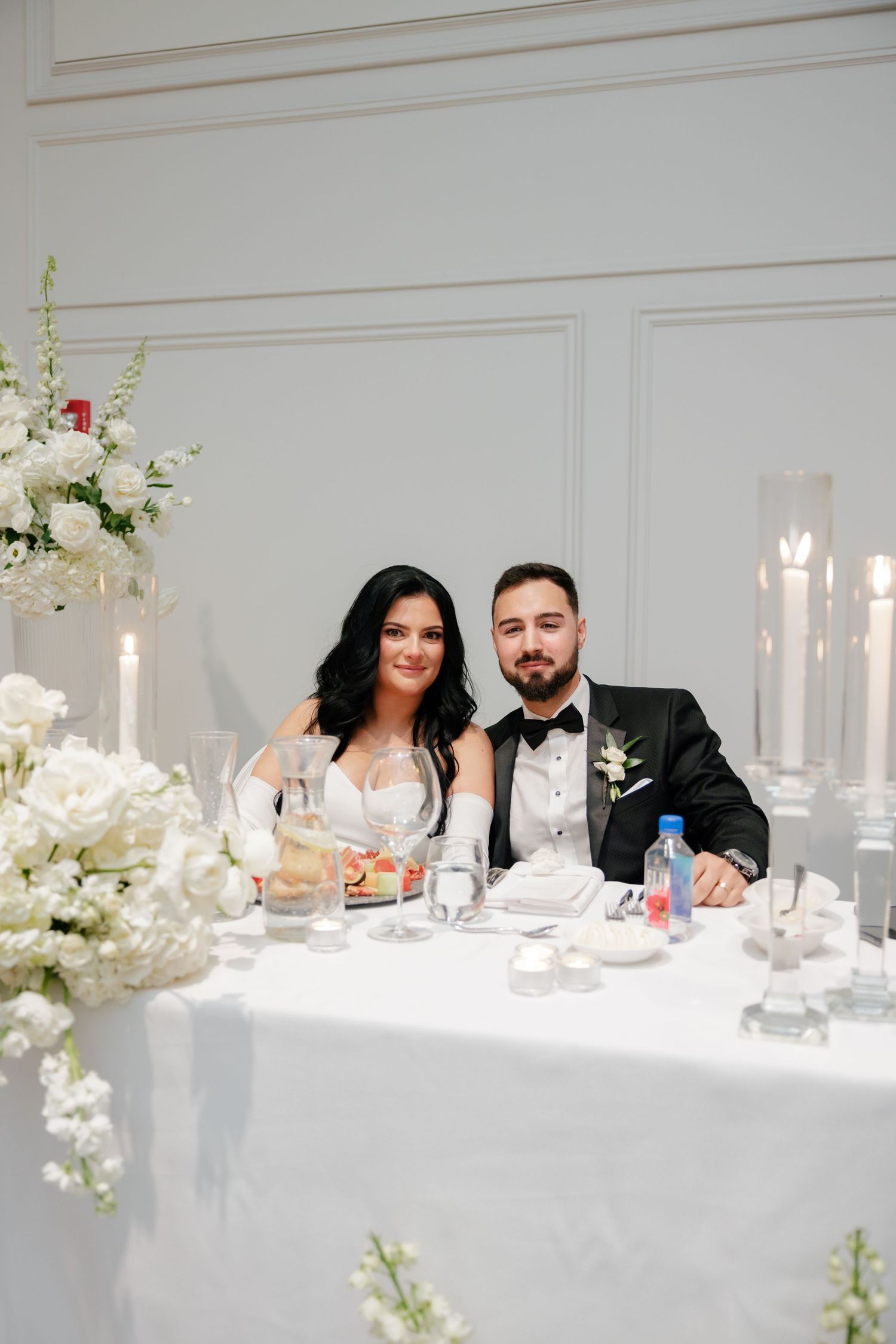 A wedding couple sits at their reception table decorated with white floral arrangements and candles.