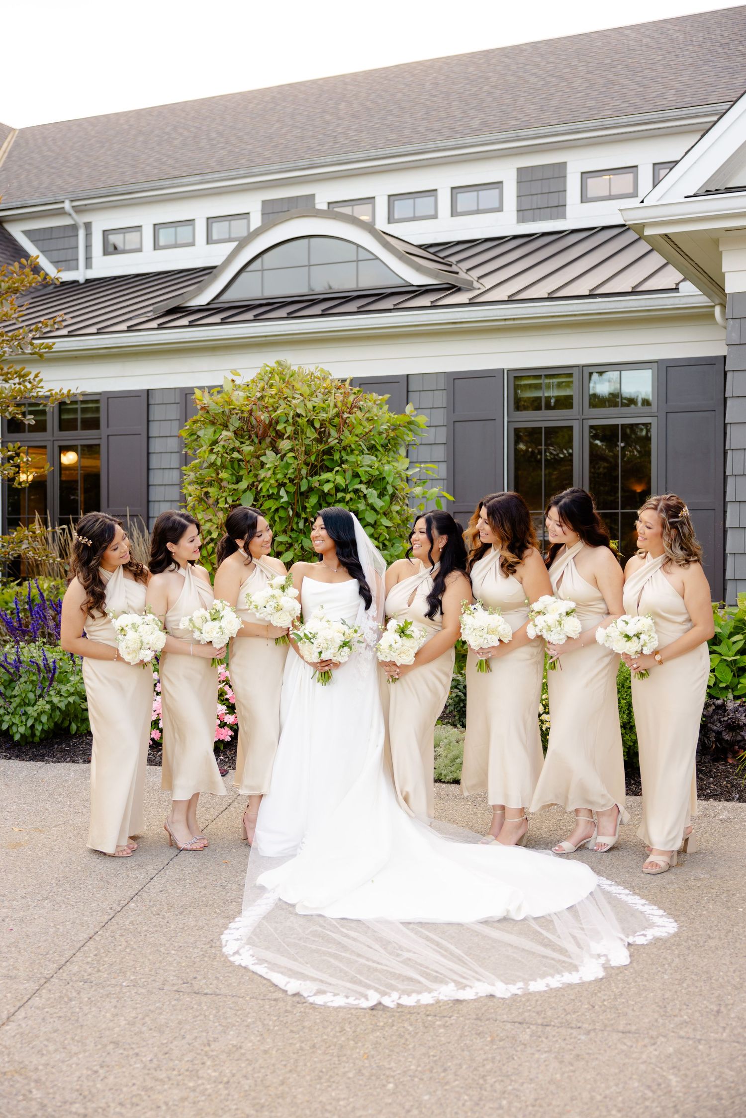 A bridal party gathers in front of a modern building wearing matching cream-colored dresses.