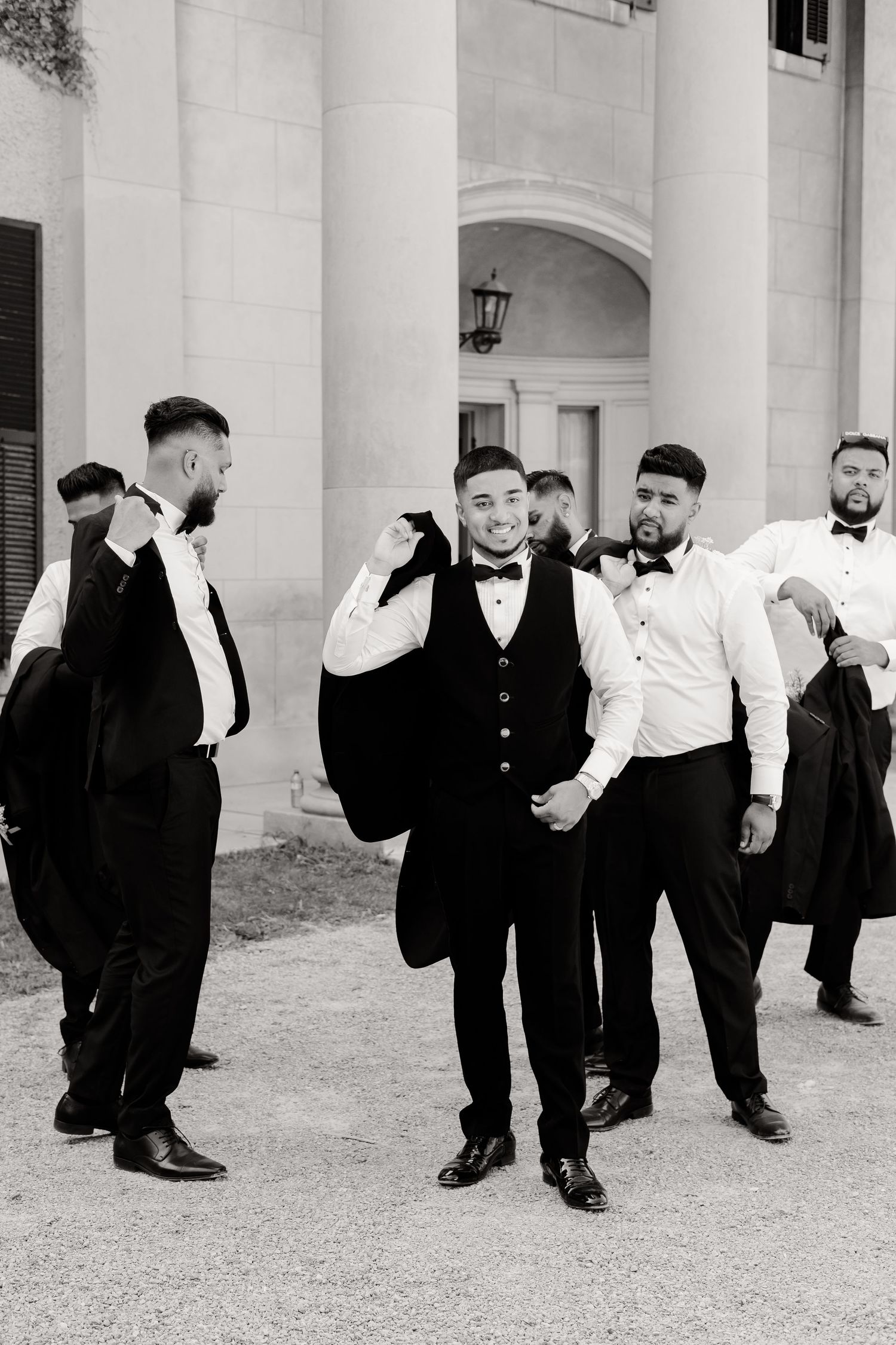 A group of groomsmen in formal attire share a lighthearted moment outside a white pillared building.