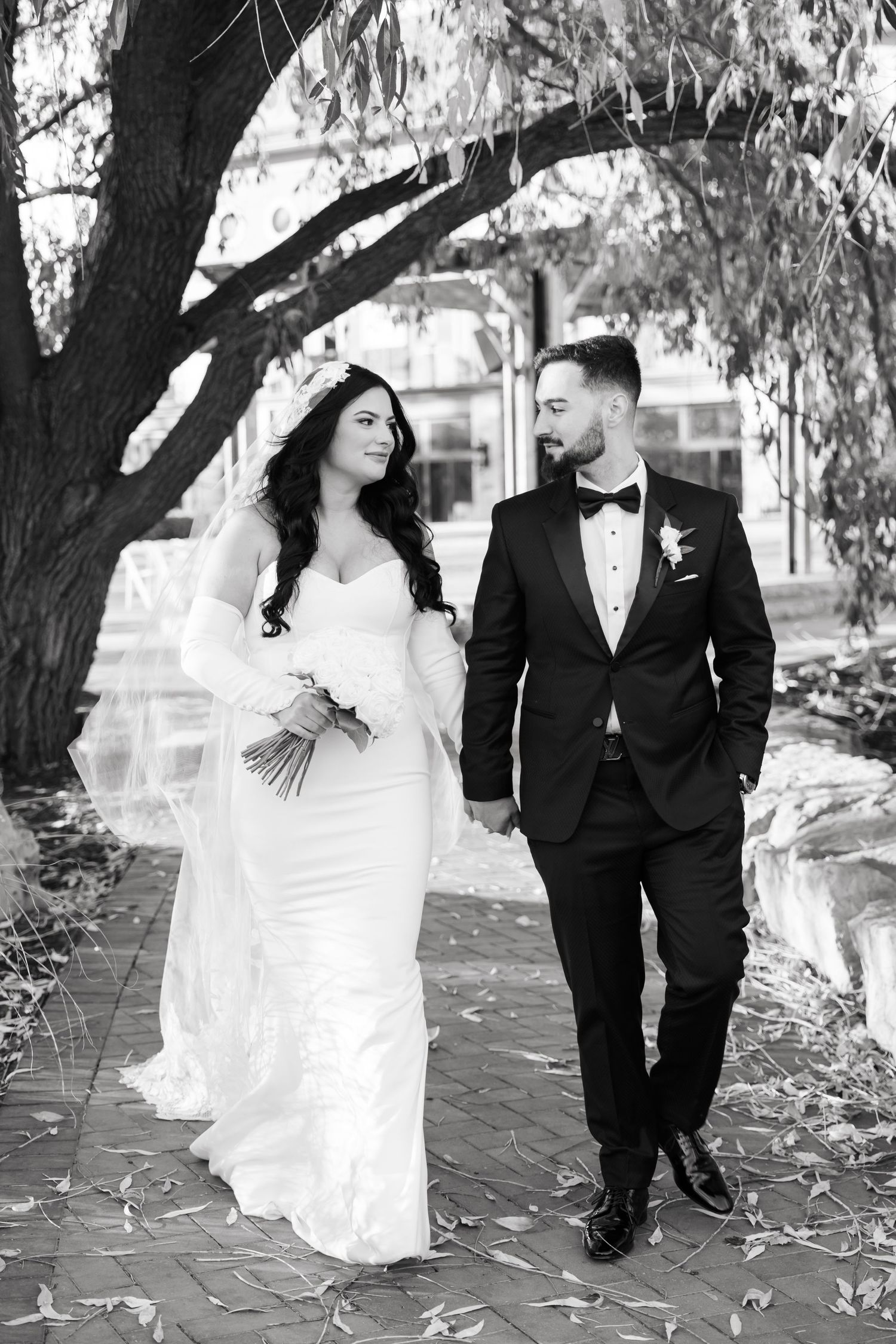 Black and white wedding portrait of a couple walking beneath a dramatic tree canopy.