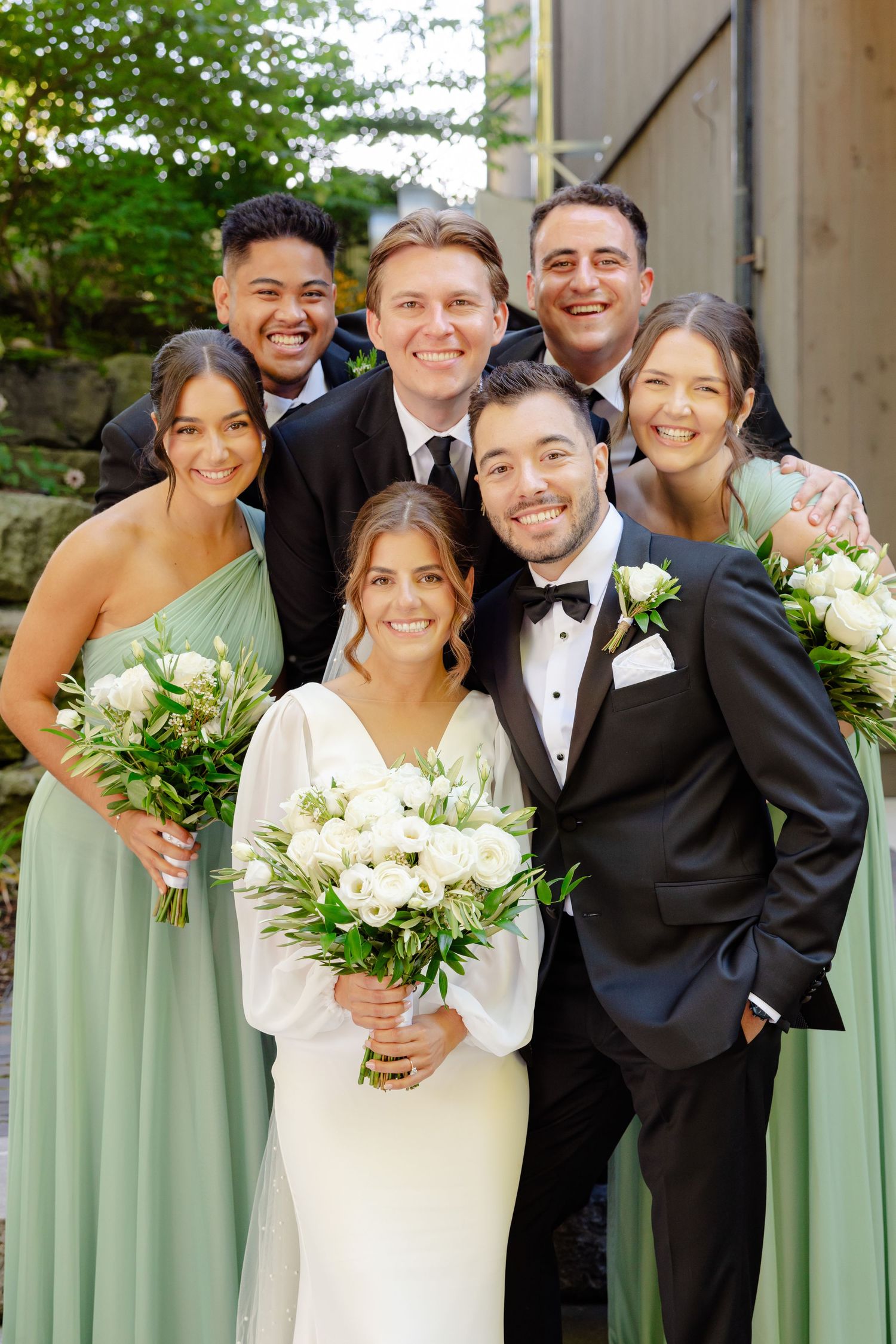 Wedding party in sage green bridesmaid dresses and black tuxedos pose together with white floral bouquets outdoors.