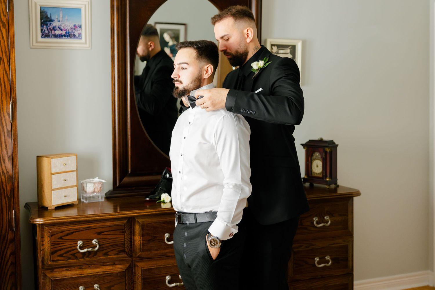 Two people getting ready in formal attire next to an antique dresser with oval mirror.