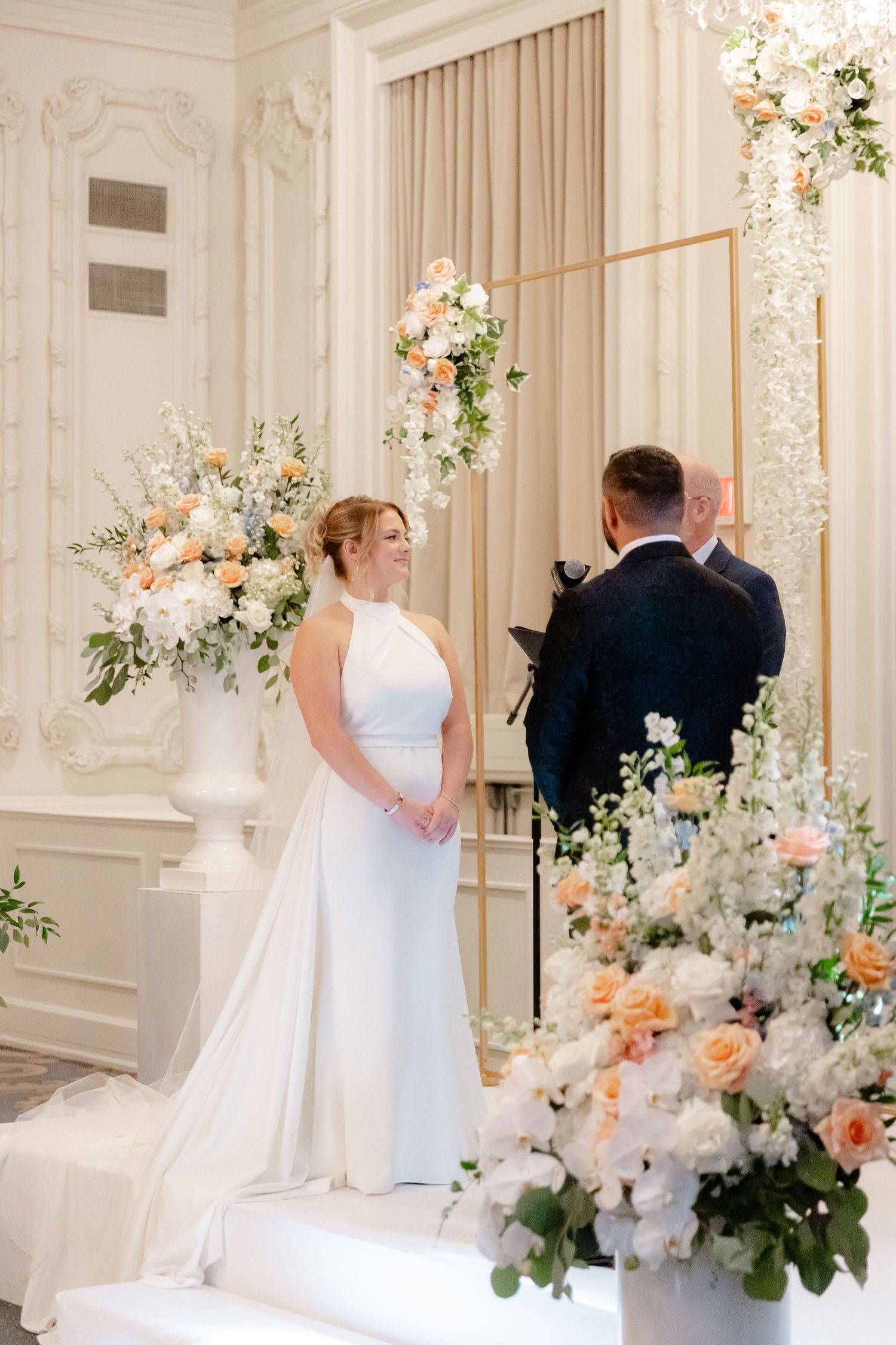 Elegant wedding ceremony setup with white floral arrangements and delicate peach accents in an ornate ballroom.