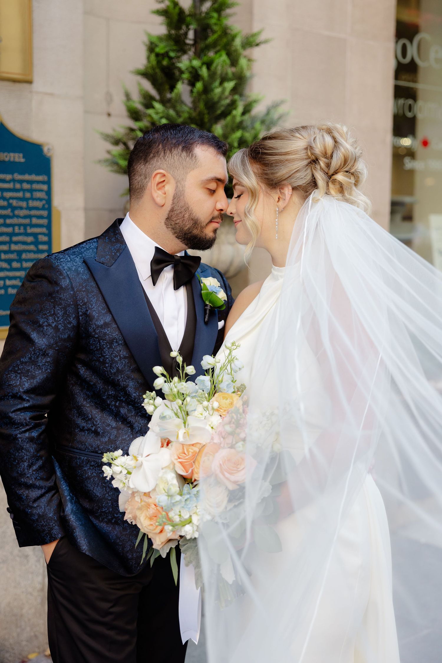 Wedding couple embraces while holding a bouquet of peach and white flowers near green topiaries.