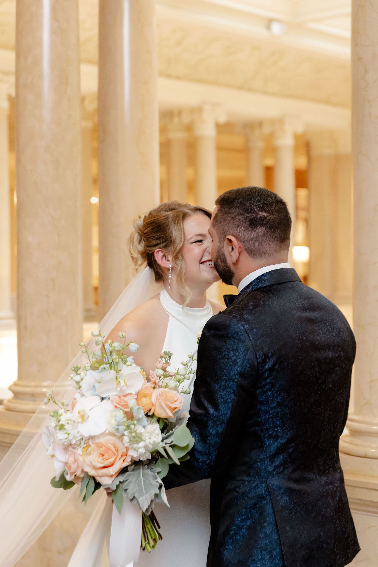 A romantic wedding moment captured in an elegant columned venue with soft lighting and peach floral bouquet.