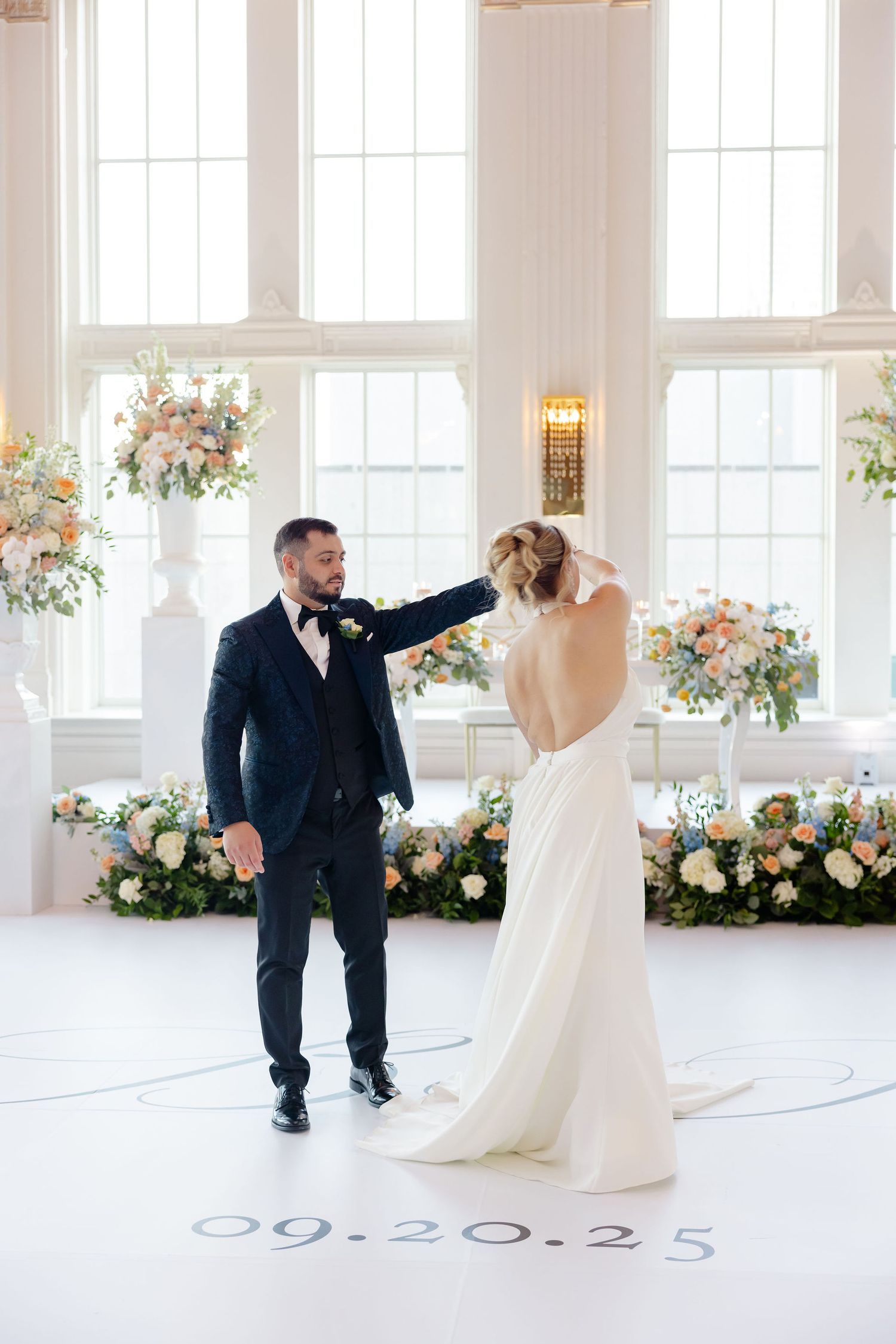 Wedding couple dancing and twirling in an elegant white ballroom with large windows and floral decorations.
