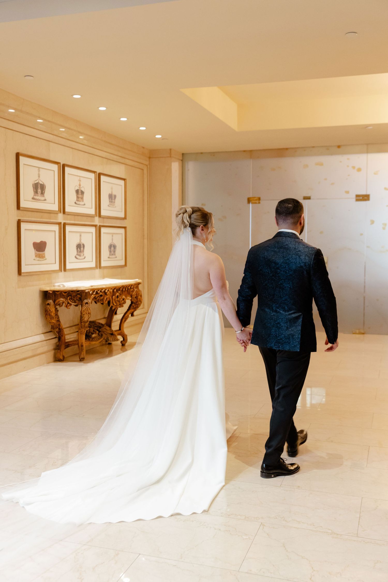 Wedding couple walks down an elegant hallway with framed artwork and warm lighting.