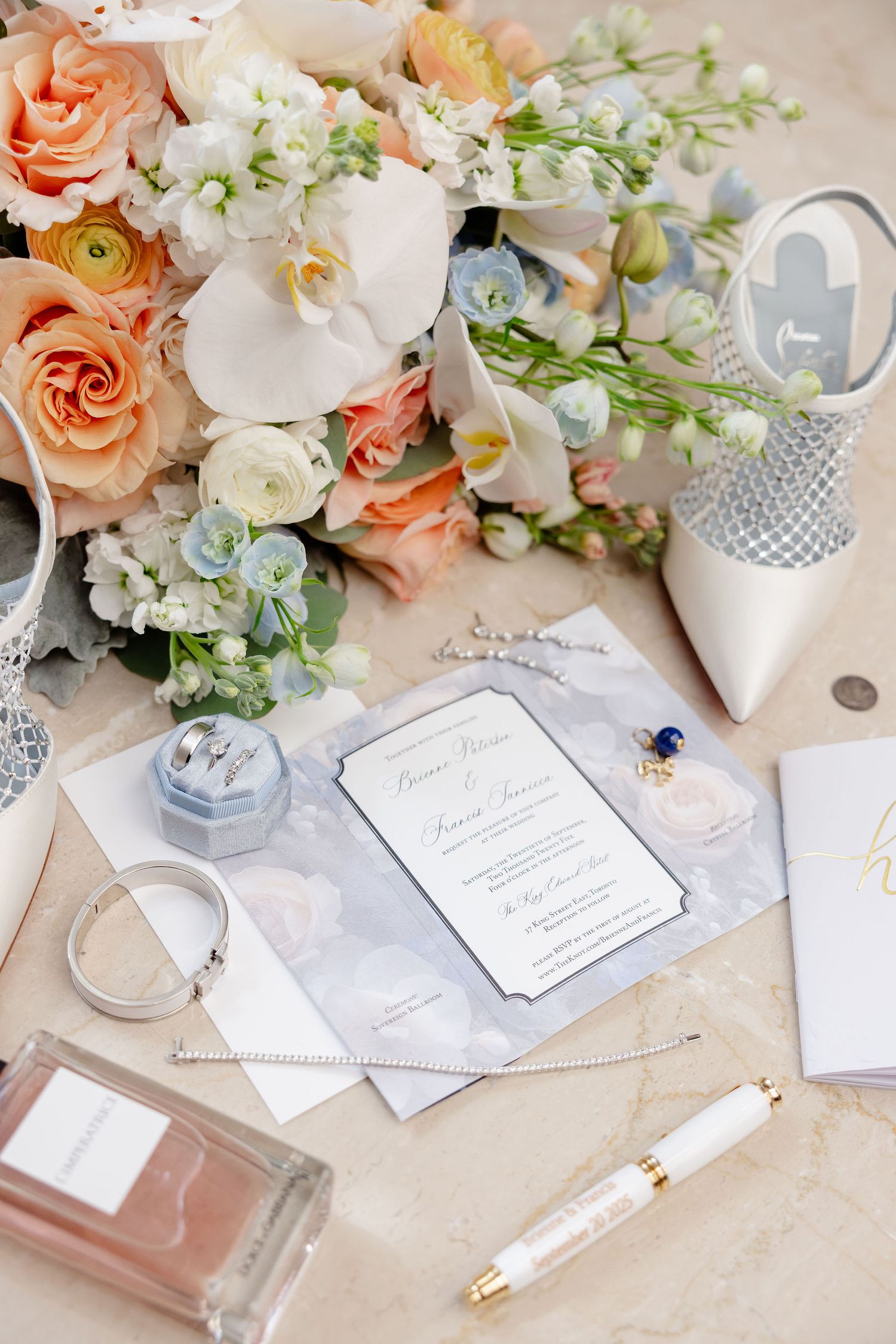Wedding details flat lay with peach and white flowers, invitations, perfume and bridal accessories on marble surface.
