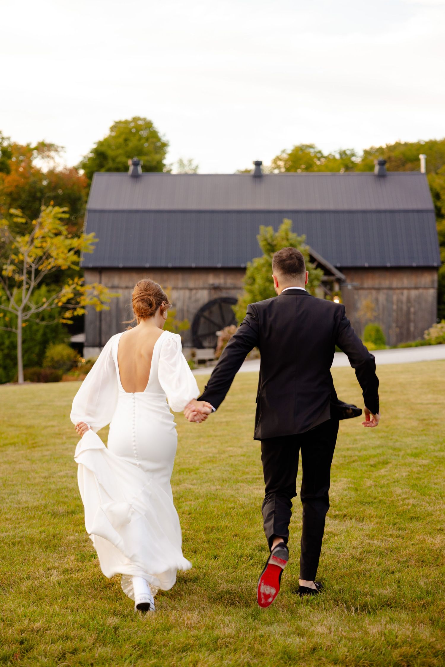 A couple running hand in hand across a grassy field towards a rustic barn.