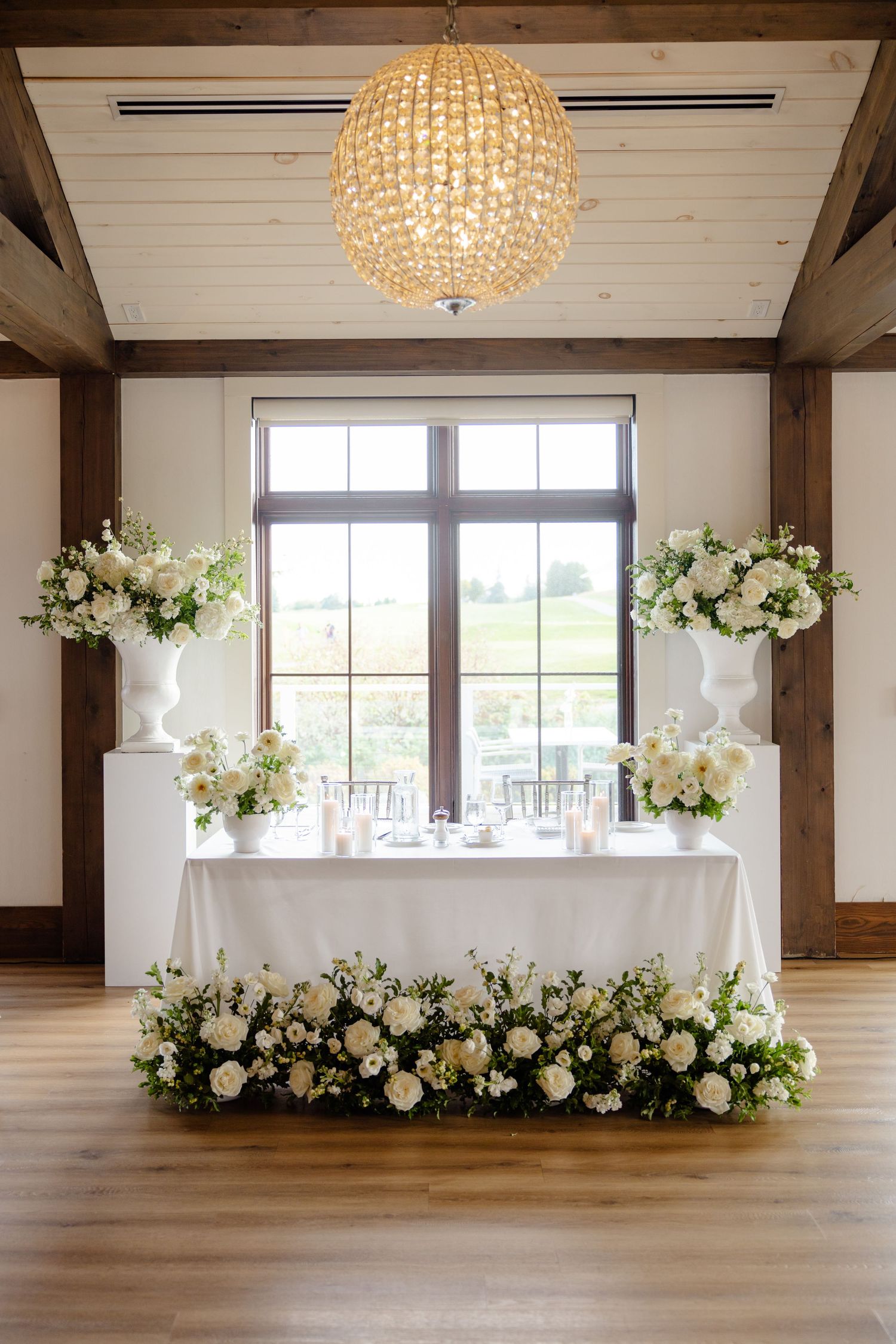 Elegant wedding reception table with crystal chandelier and white floral arrangements framing a large window.
