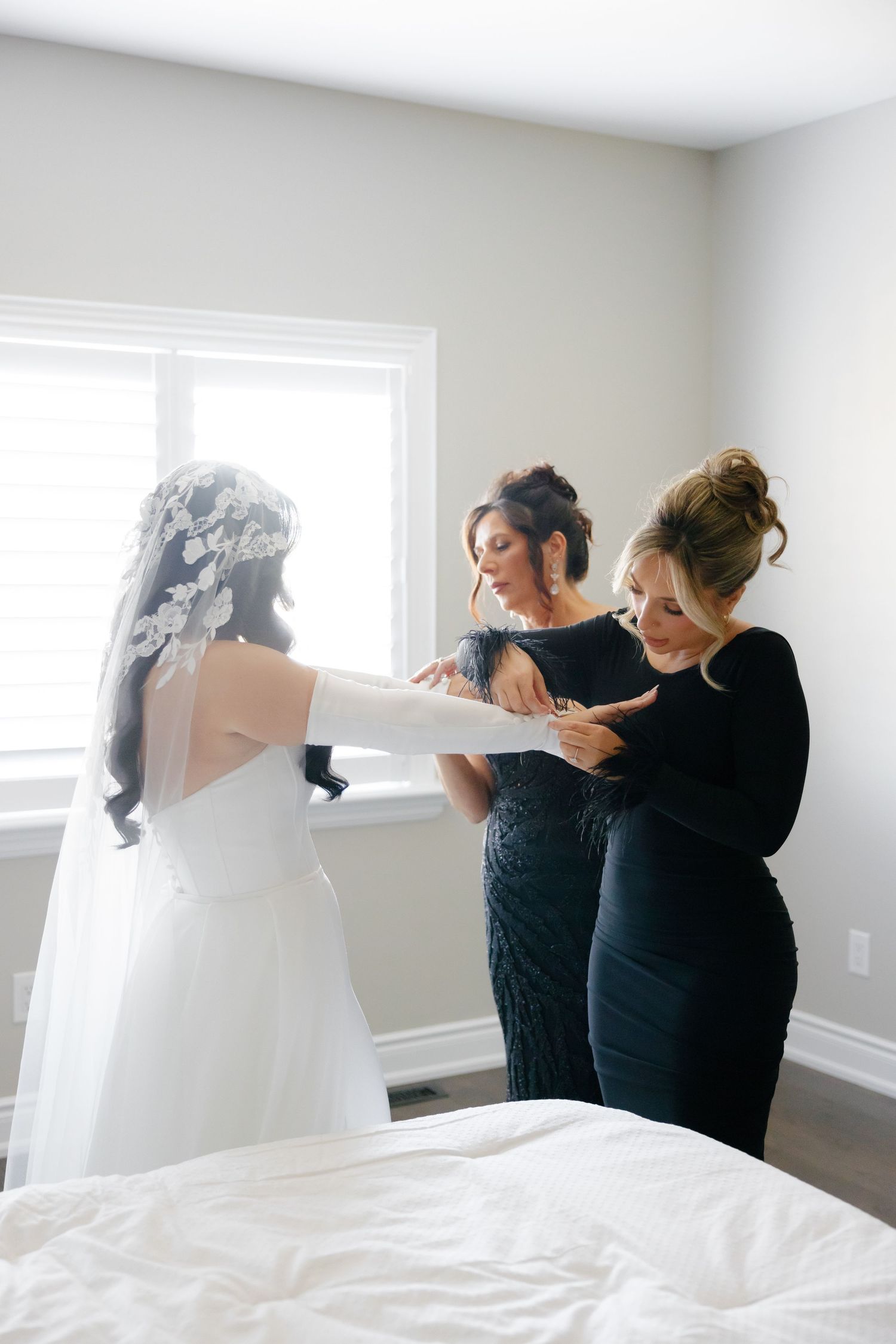 Two attendants in black dresses help arrange a bride's wedding gown by a sunny window.