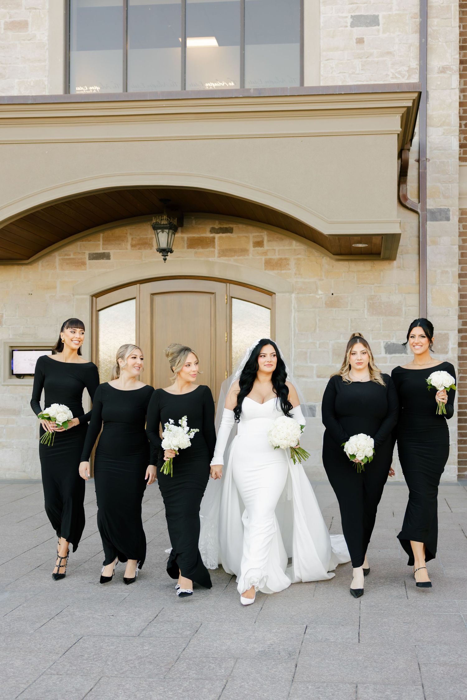 Bridal party in elegant black dresses stands with bride in white gown under arched entrance.