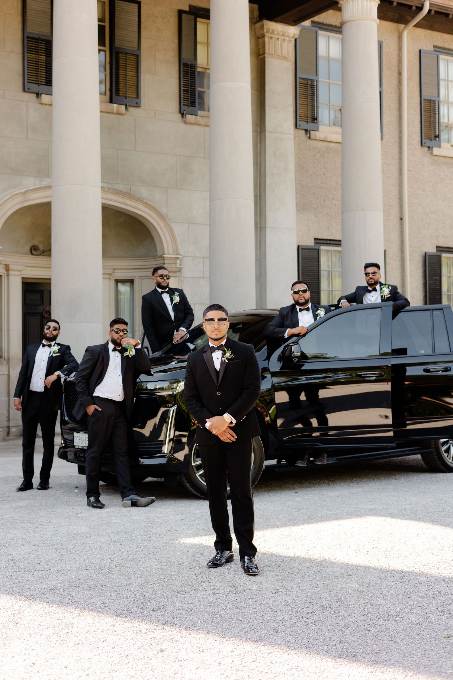 Wedding party members in formal attire gather around a black SUV outside an elegant building with columns.