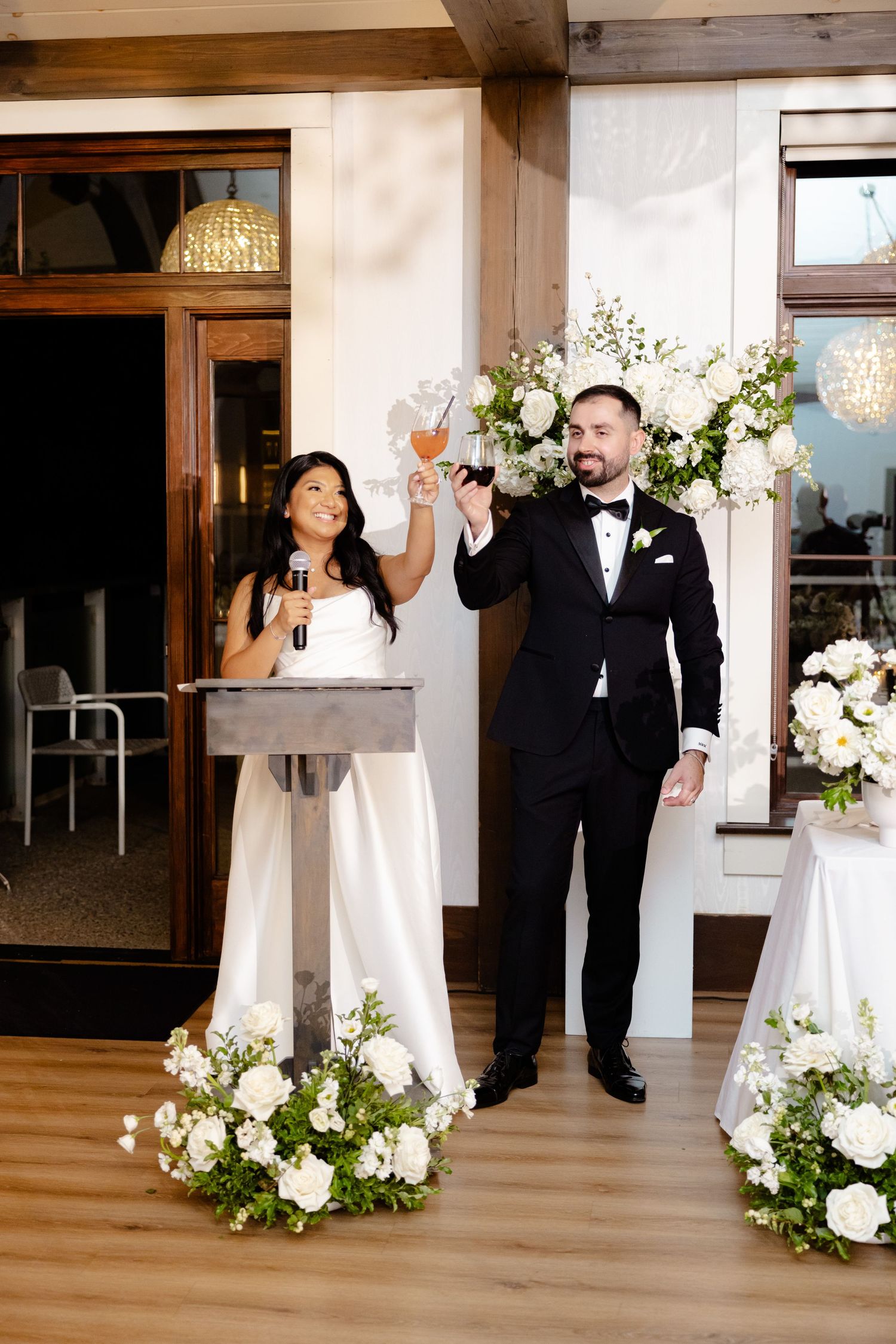 Newly married couple toasts with champagne during wedding reception surrounded by elegant white floral arrangements.