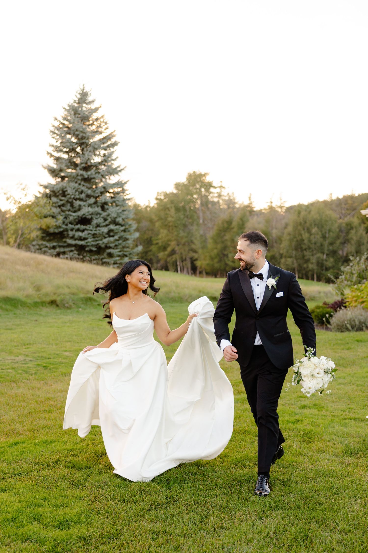 Happy couple running through garden grounds with flowing white wedding dress and bouquet.