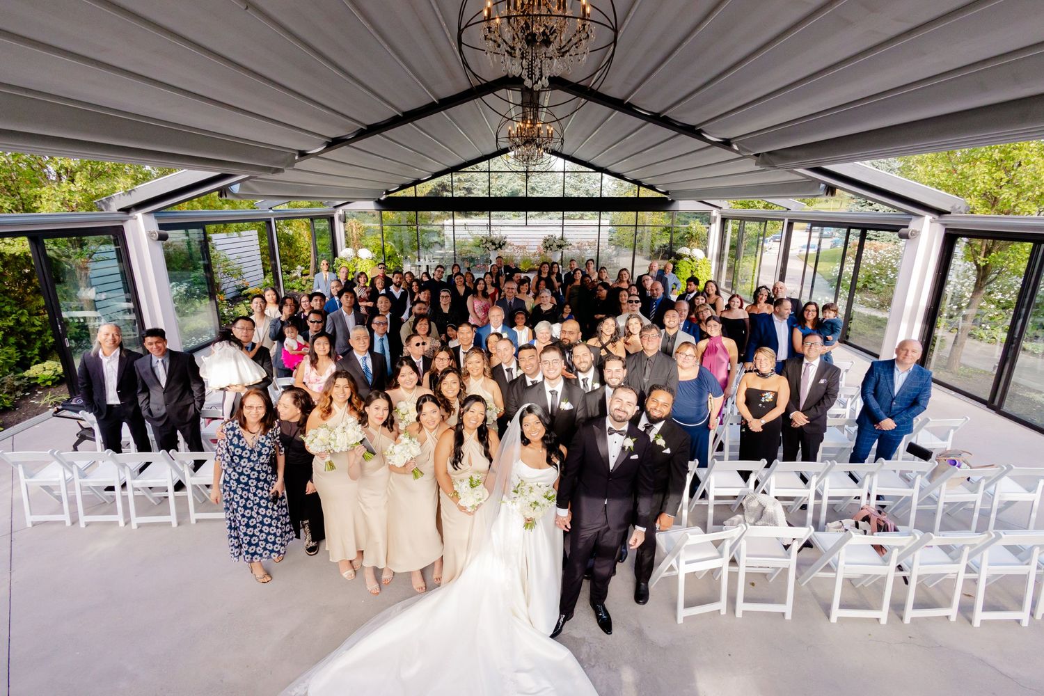 Large group wedding photo showing all guests and wedding party in an elegant glass-enclosed venue.