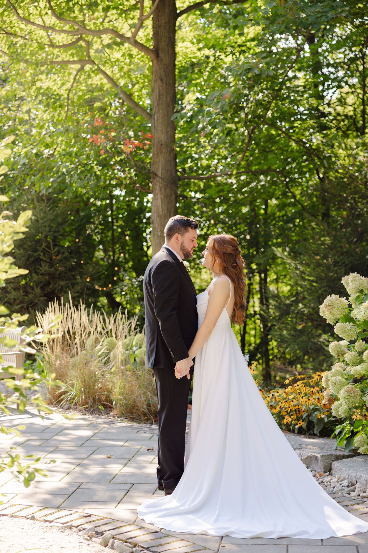 A romantic wedding portrait on a garden path with hydrangeas and sunlit trees in the background.