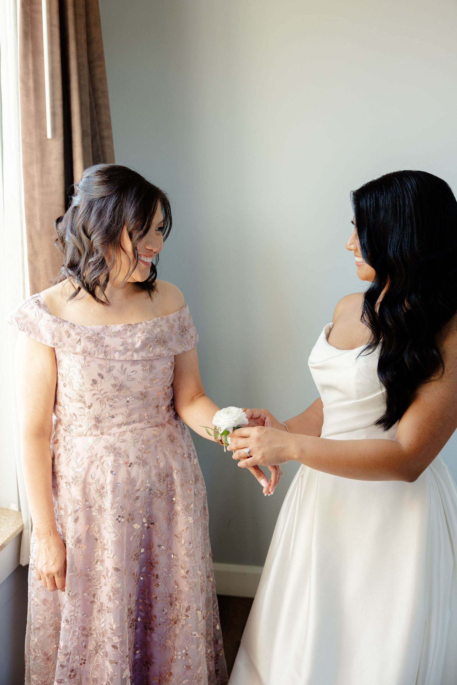 Two people in complementary formal dresses share a moment by a window during wedding preparations.