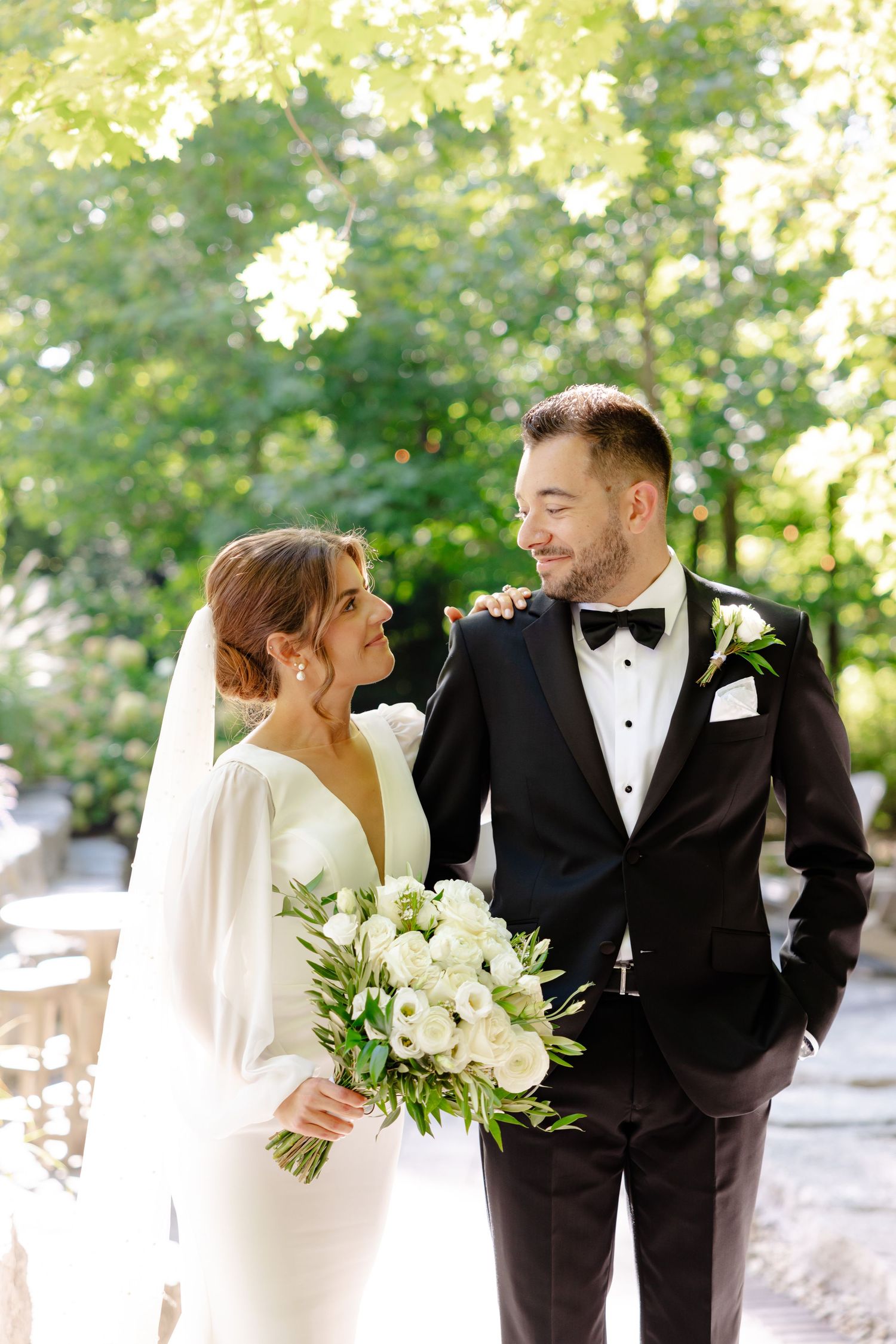 Candid wedding photo showing natural laughter between couple with white floral bouquet under tree canopy.