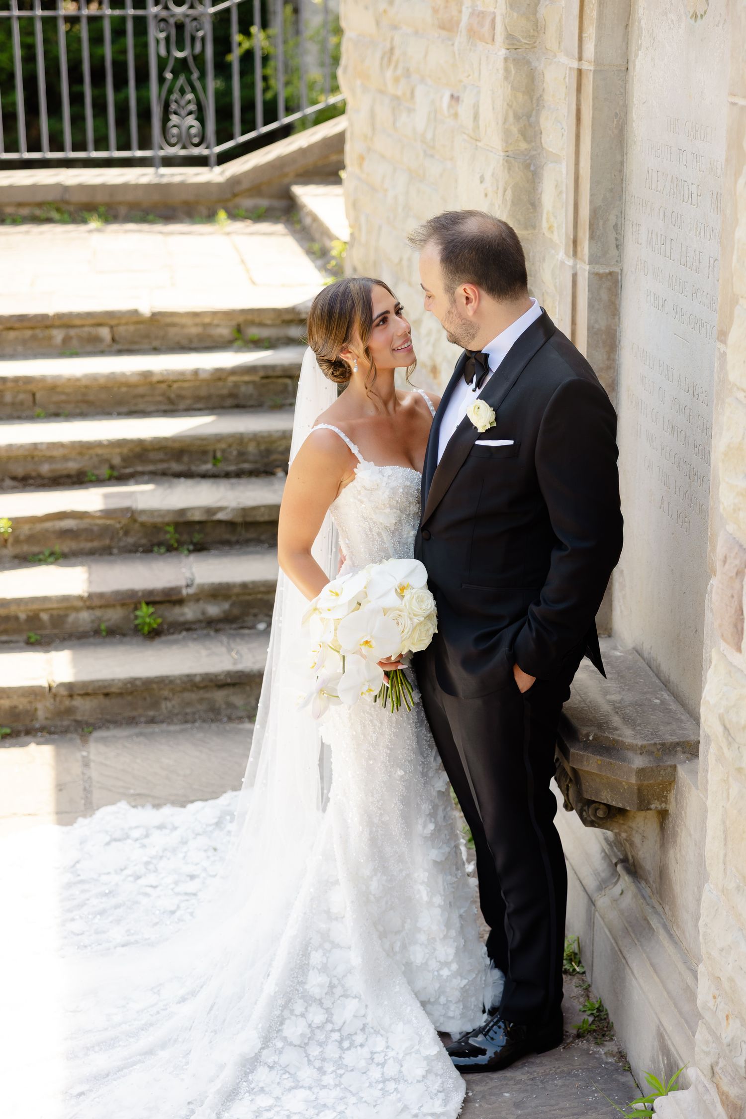 A romantic wedding portrait shows a couple standing together by stone stairs, with the bride holding a white bouquet.