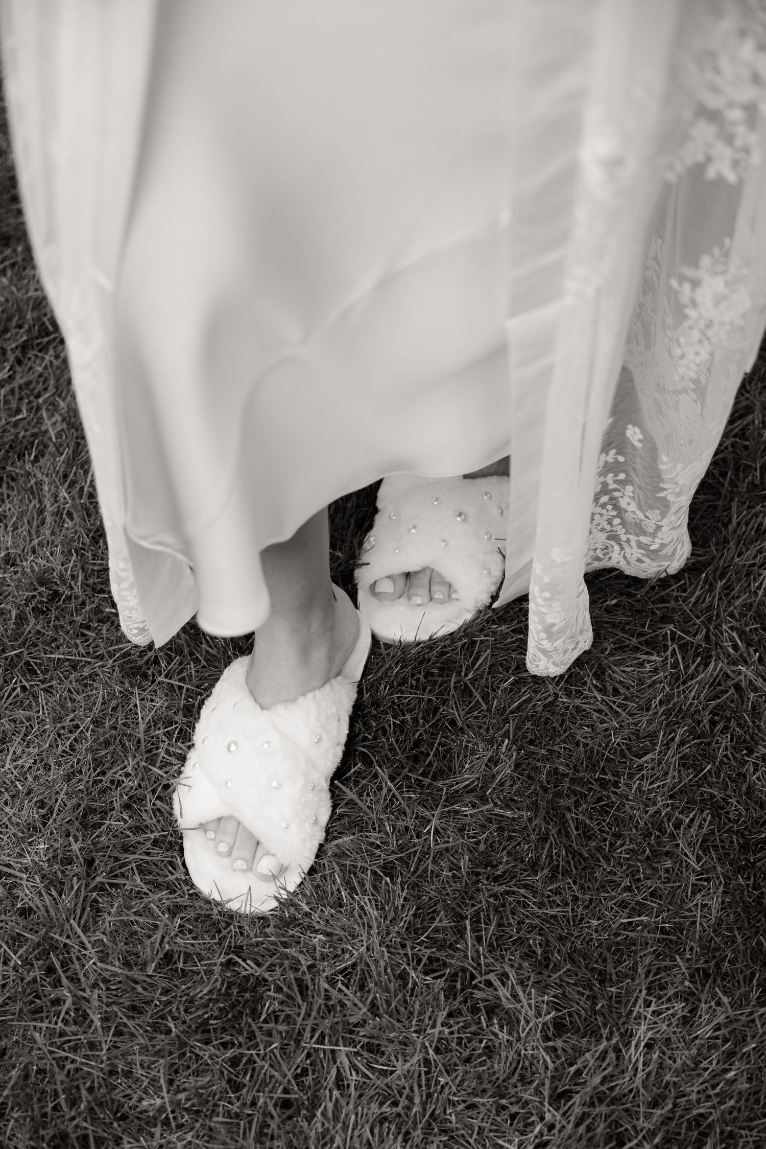 Wedding dress hem and white ballet flats visible against dark grass at an outdoor ceremony.