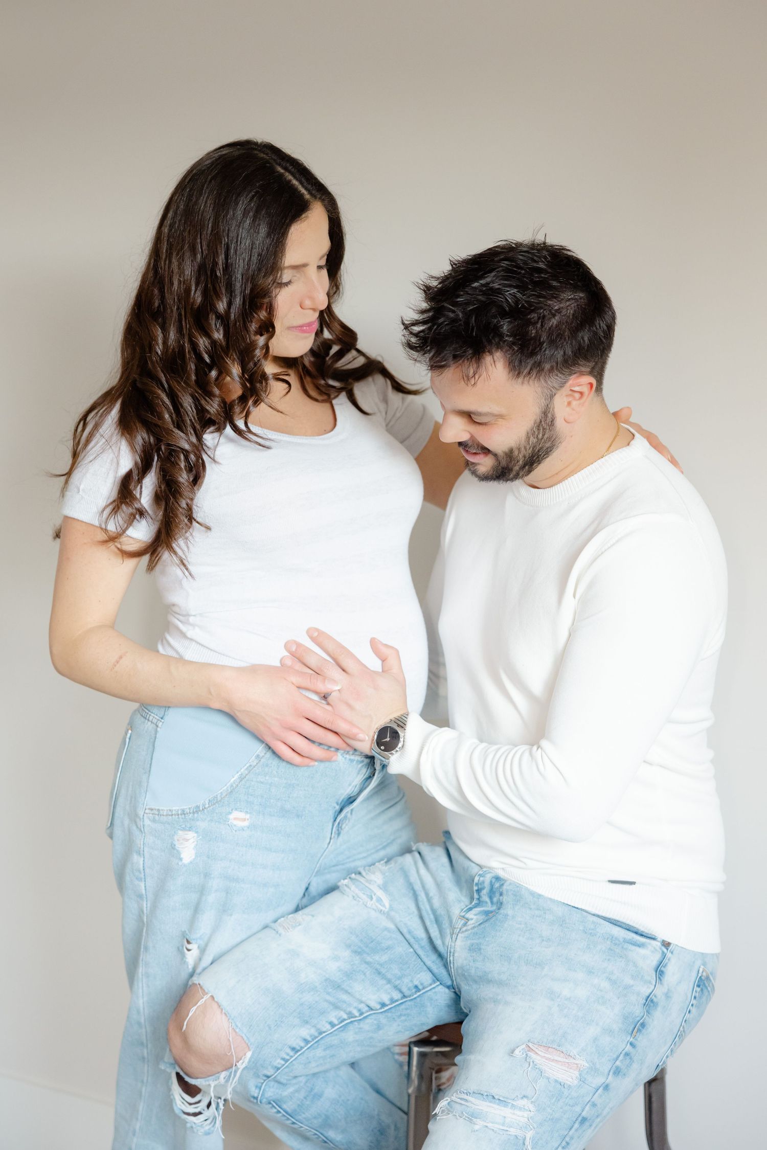 Couple in white tops and ripped jeans embrace, with the woman's pregnant belly visible as they look down tenderly.