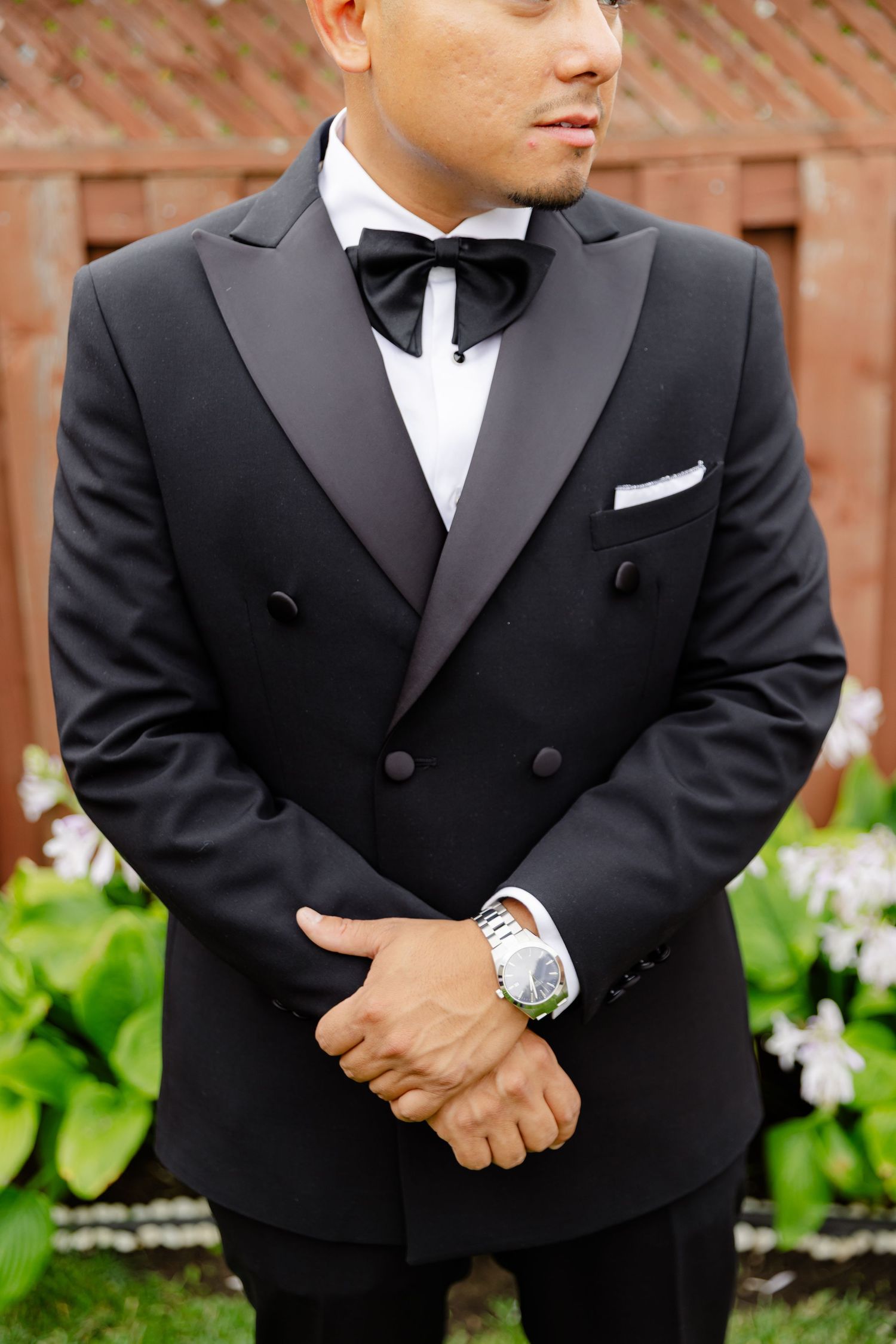 A close-up shot of a formal black double-breasted tuxedo with bow tie and white dress shirt against a garden background.