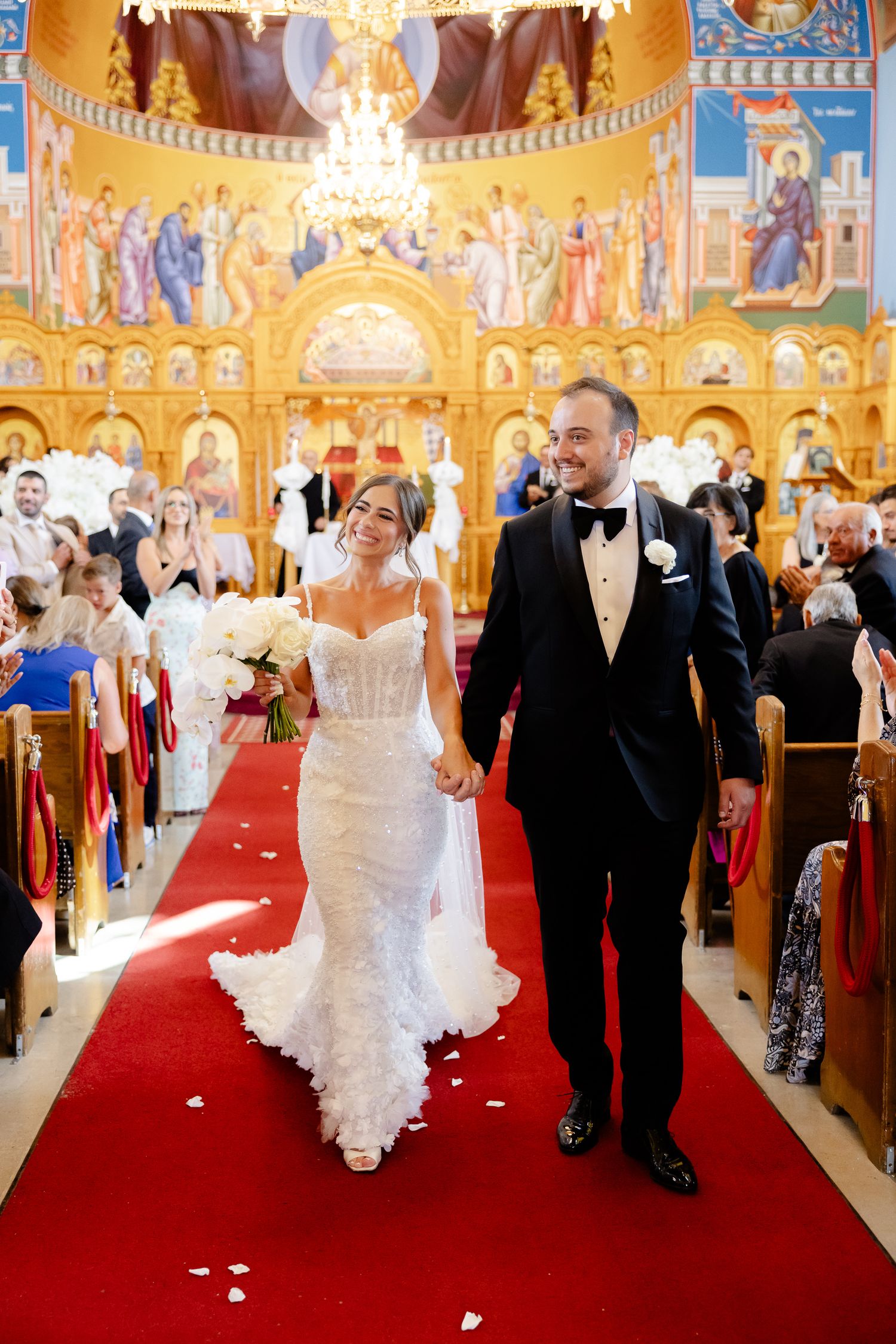 A couple walks down a red-carpeted aisle in an ornate church with golden Byzantine style murals adorning the walls.