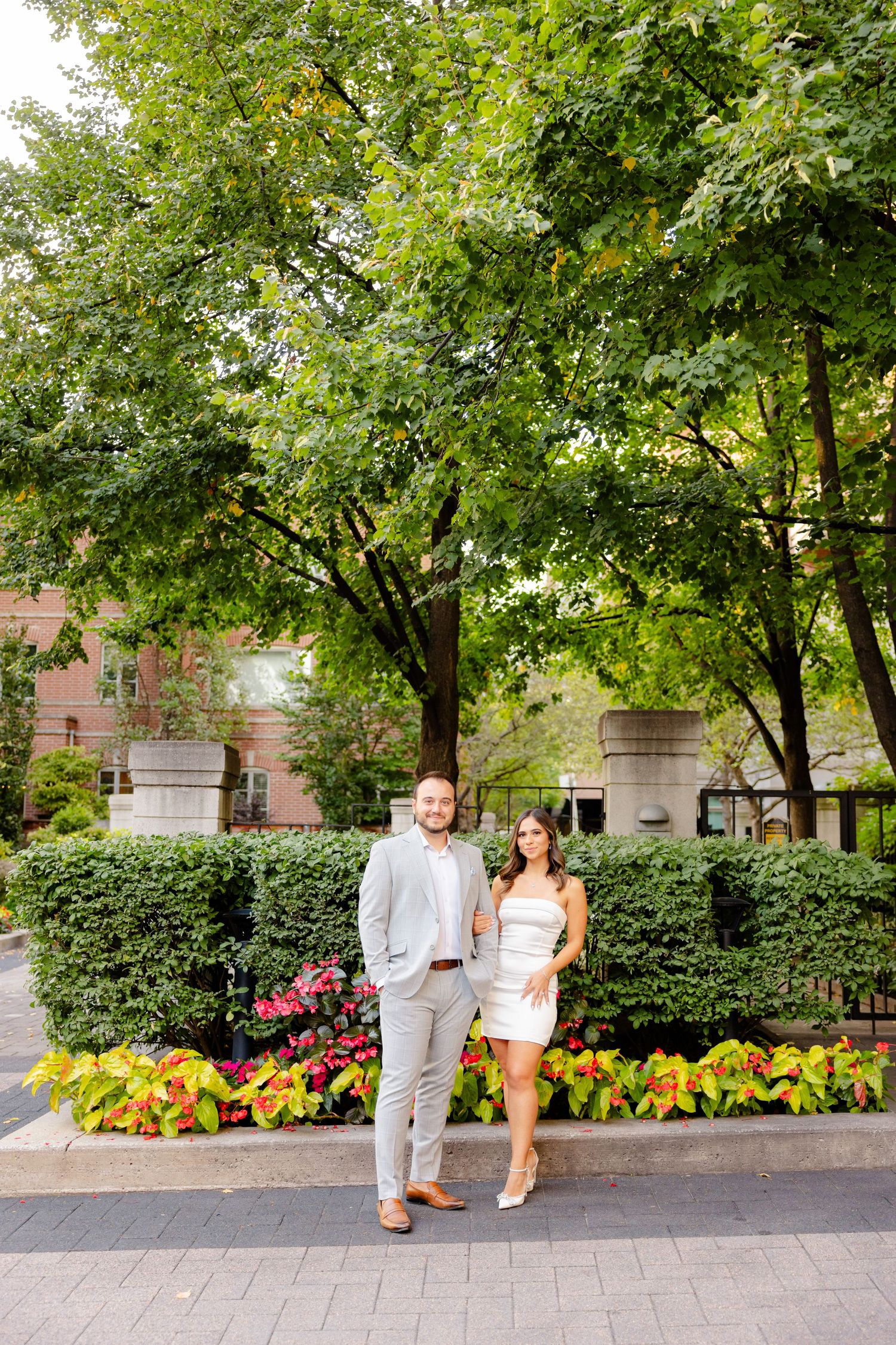 A couple in formal attire stands in a garden setting with lush greenery and colorful flowers.