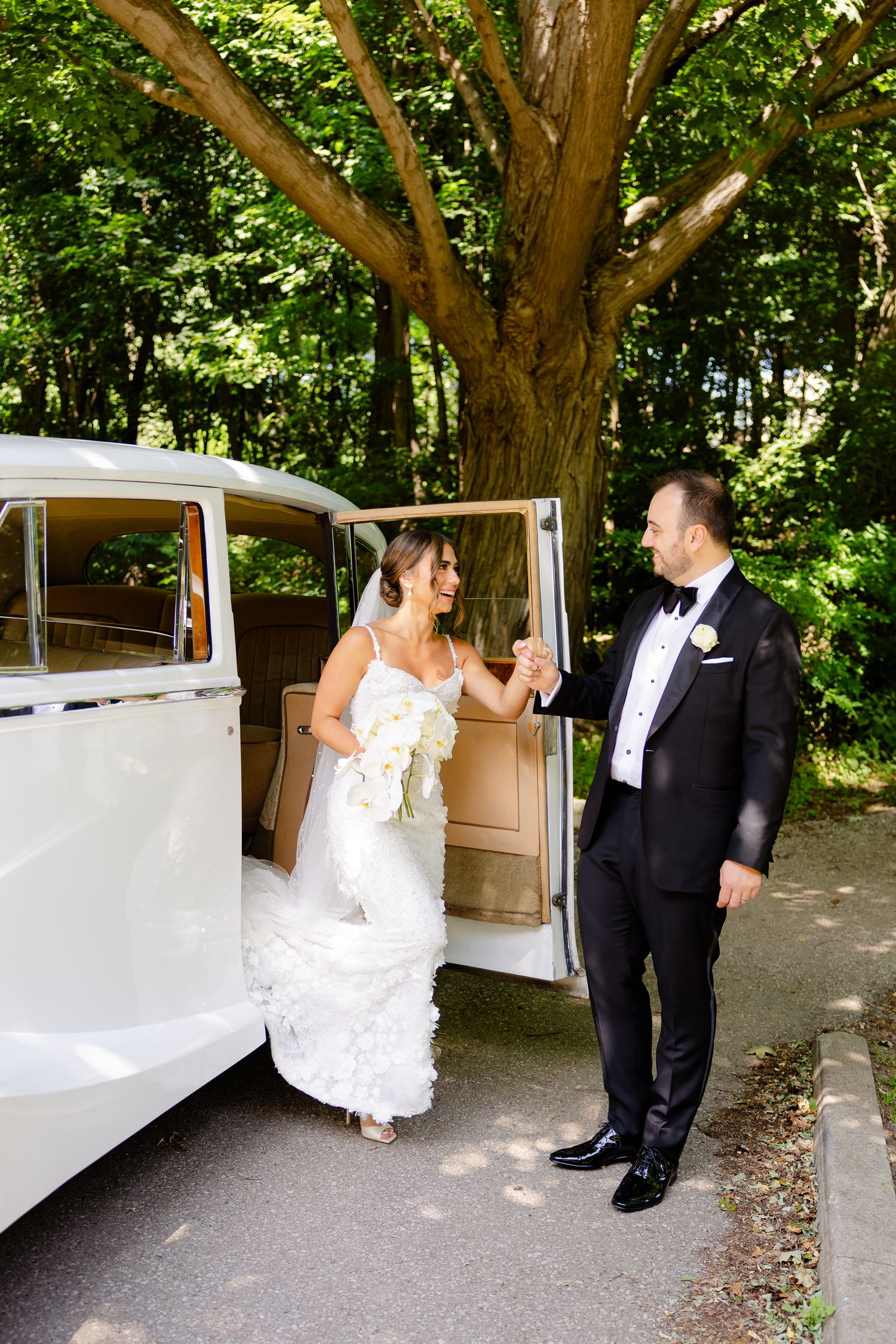 A bride and groom pose playfully with a vintage white car at their woodland wedding venue.