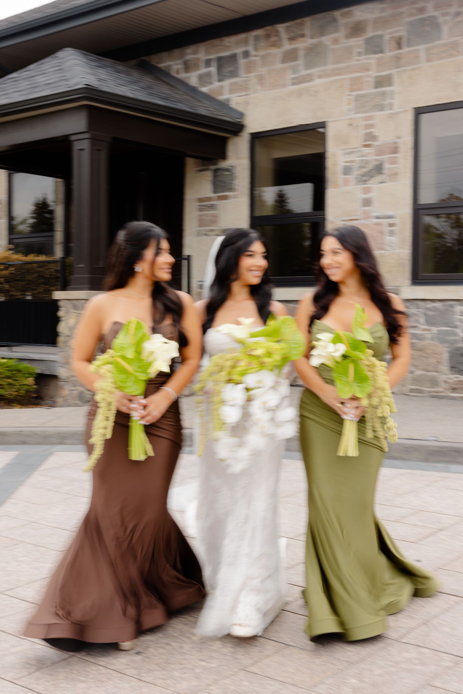 Three bridesmaids in brown and olive green dresses twirl and dance together holding bouquets outside a stone building.