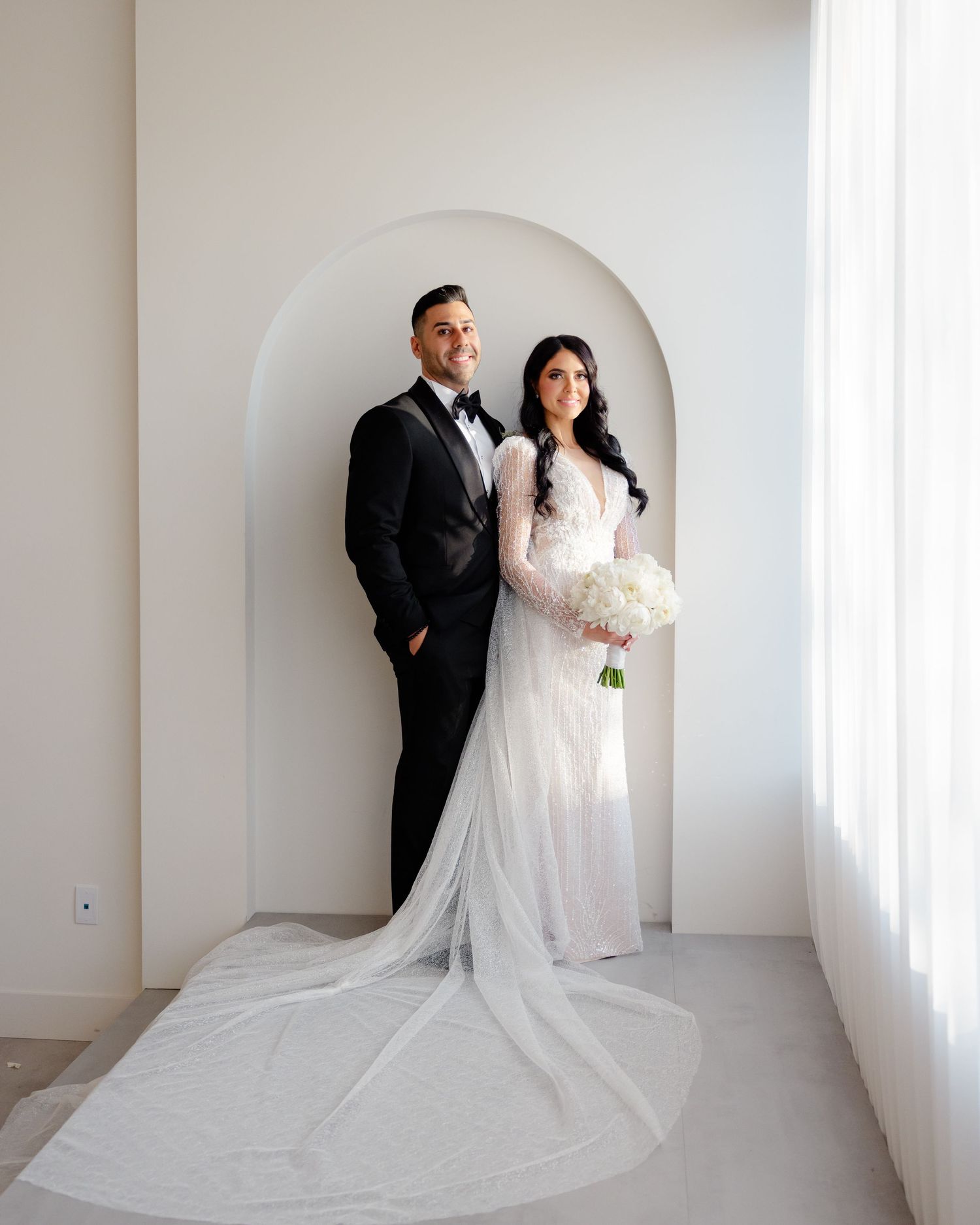 Newlyweds stand elegantly in arched alcove with bride holding white bouquet.