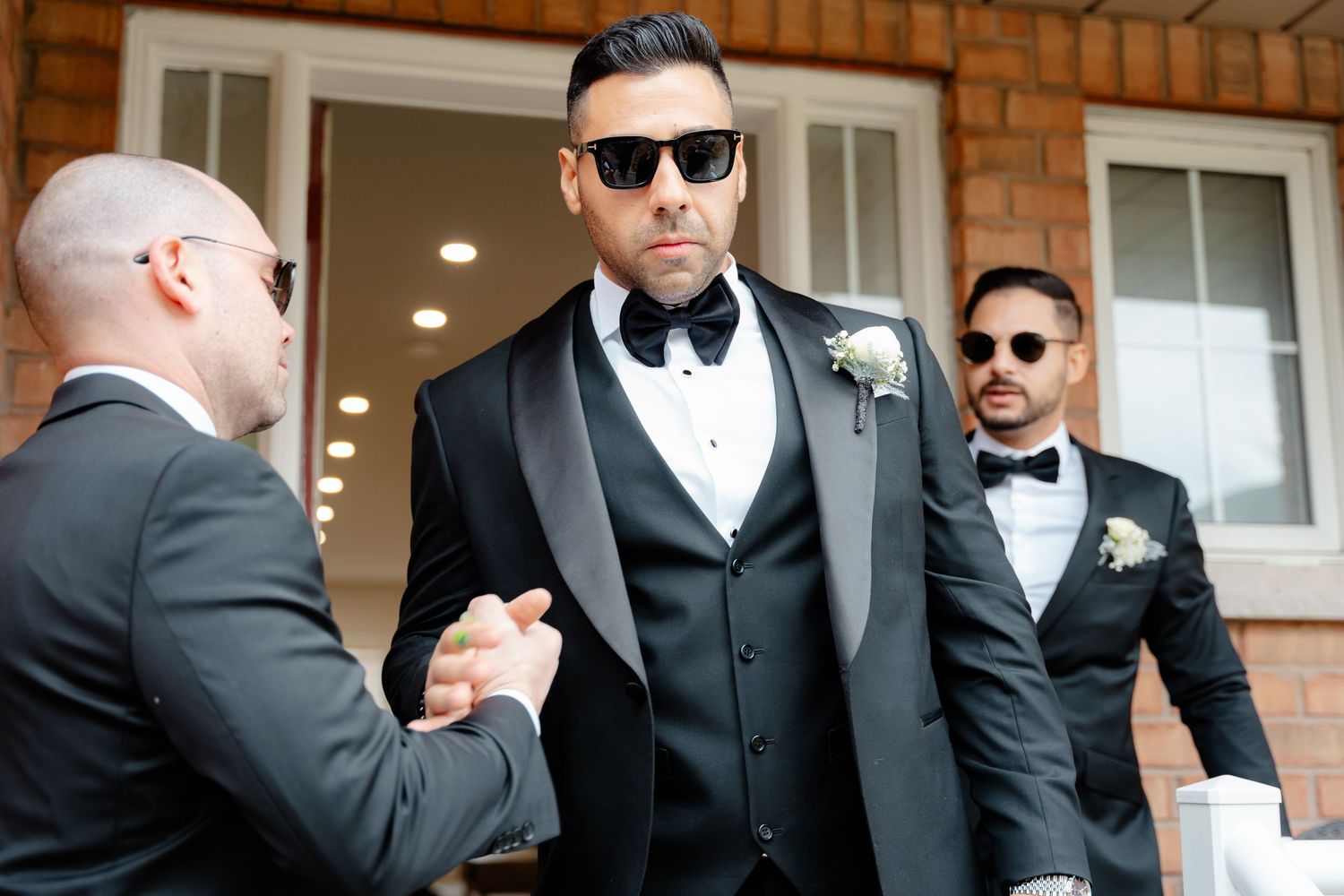 Groomsmen in black suits and sunglasses greeting each other on brick porch steps.