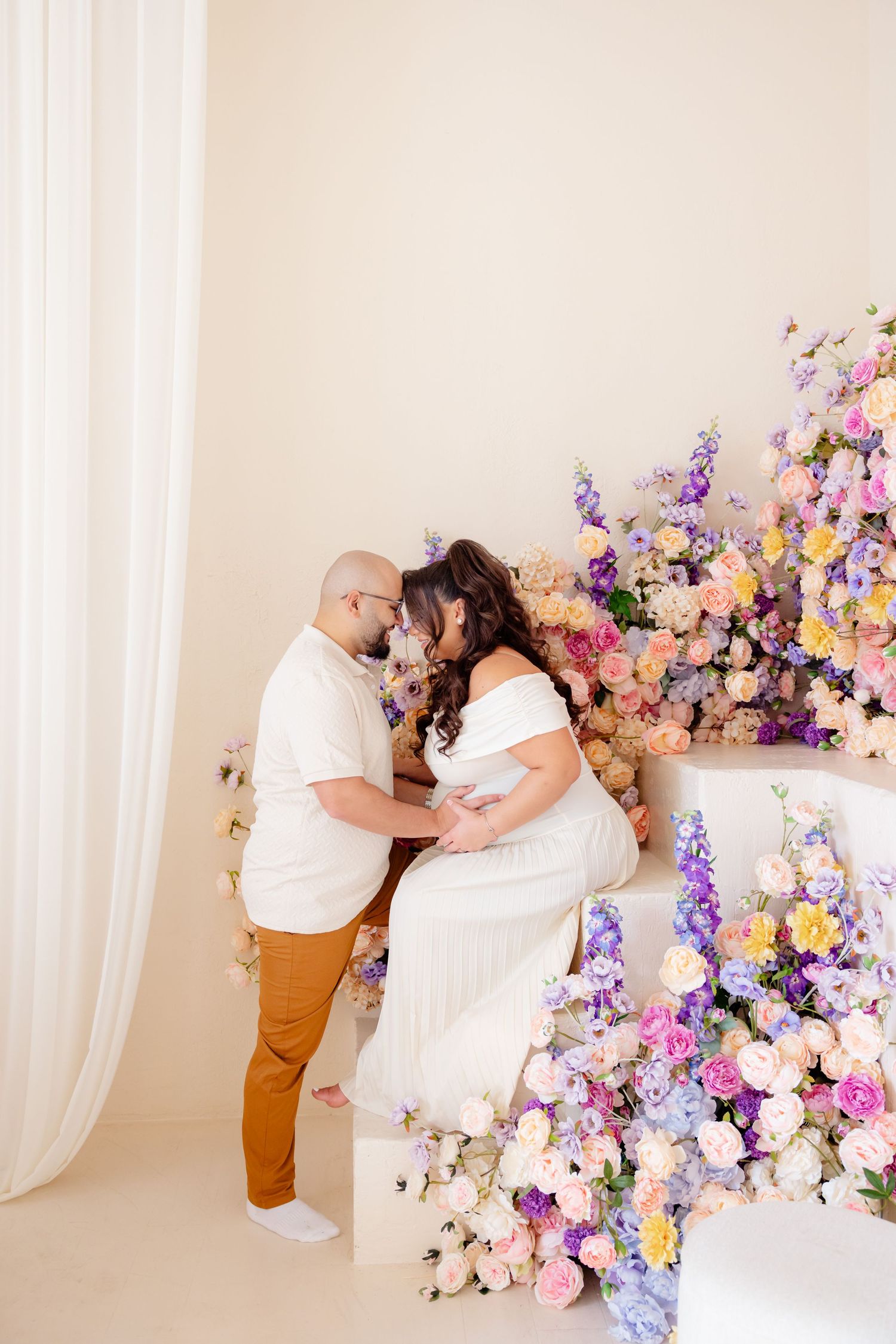 A romantic couple embraces near a beautiful floral display of pink, purple and white flowers in a bright studio setting.