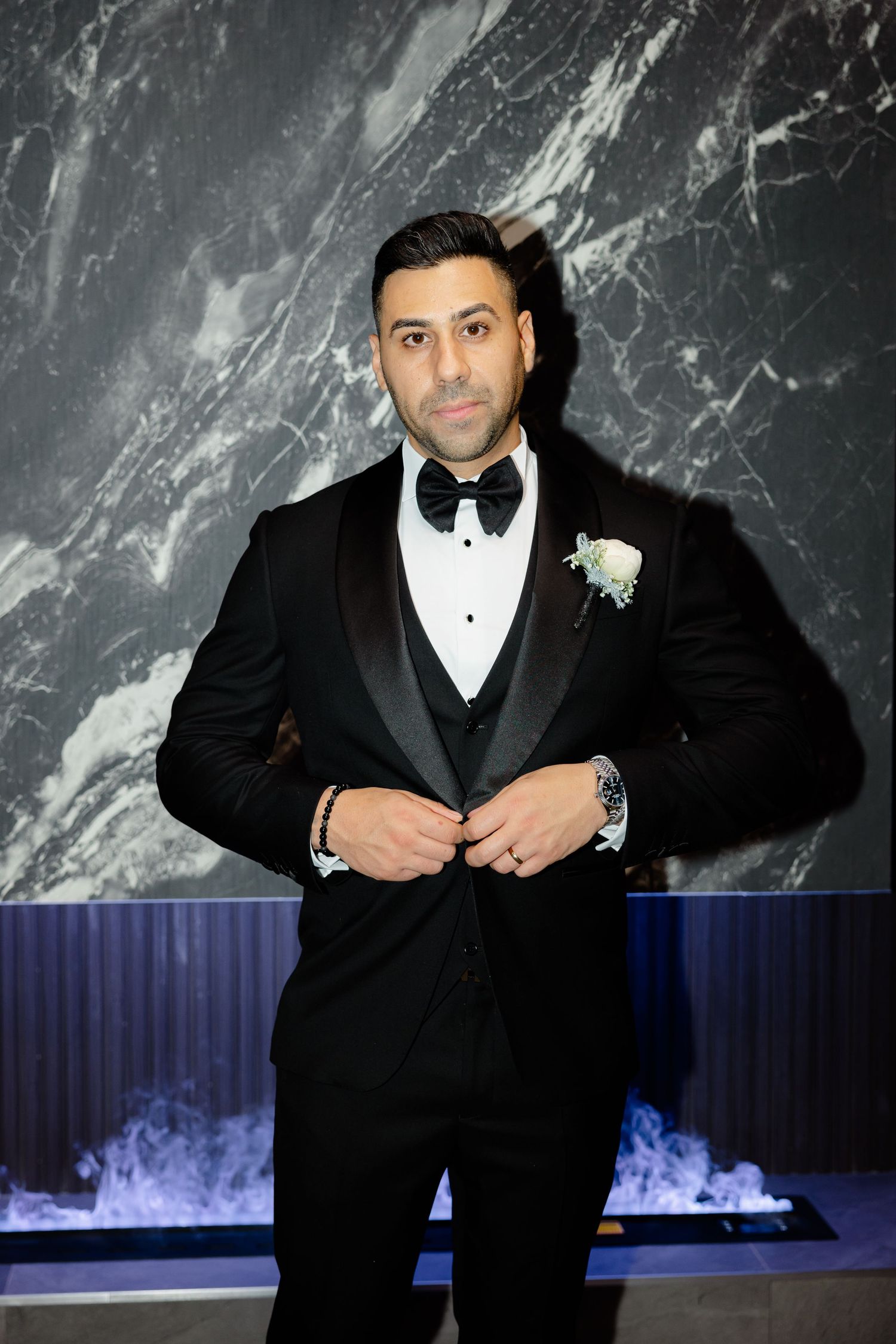 Sophisticated groom portrait in black tuxedo with white boutonniere against marble wall.