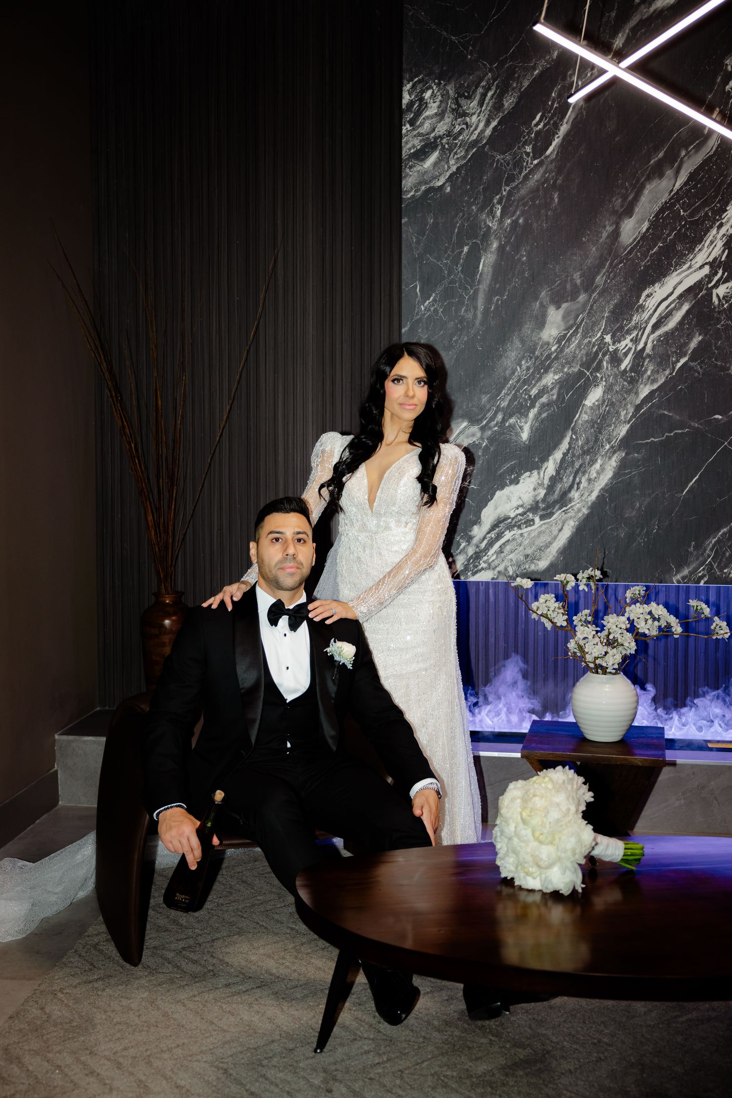 Newlywed couple poses elegantly against marble wall with bride in white lace gown and groom in black tuxedo.